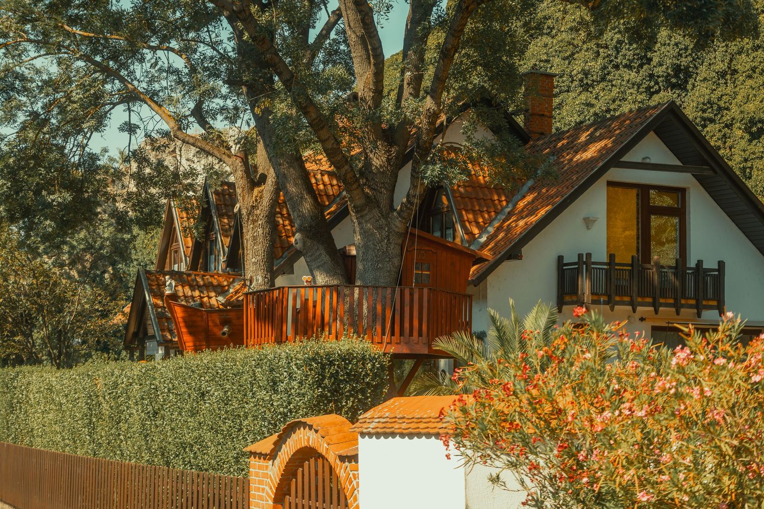 Cozy cottage with wooden deck surrounded by large oak trees and colorful flowering shrubs behind a fence.