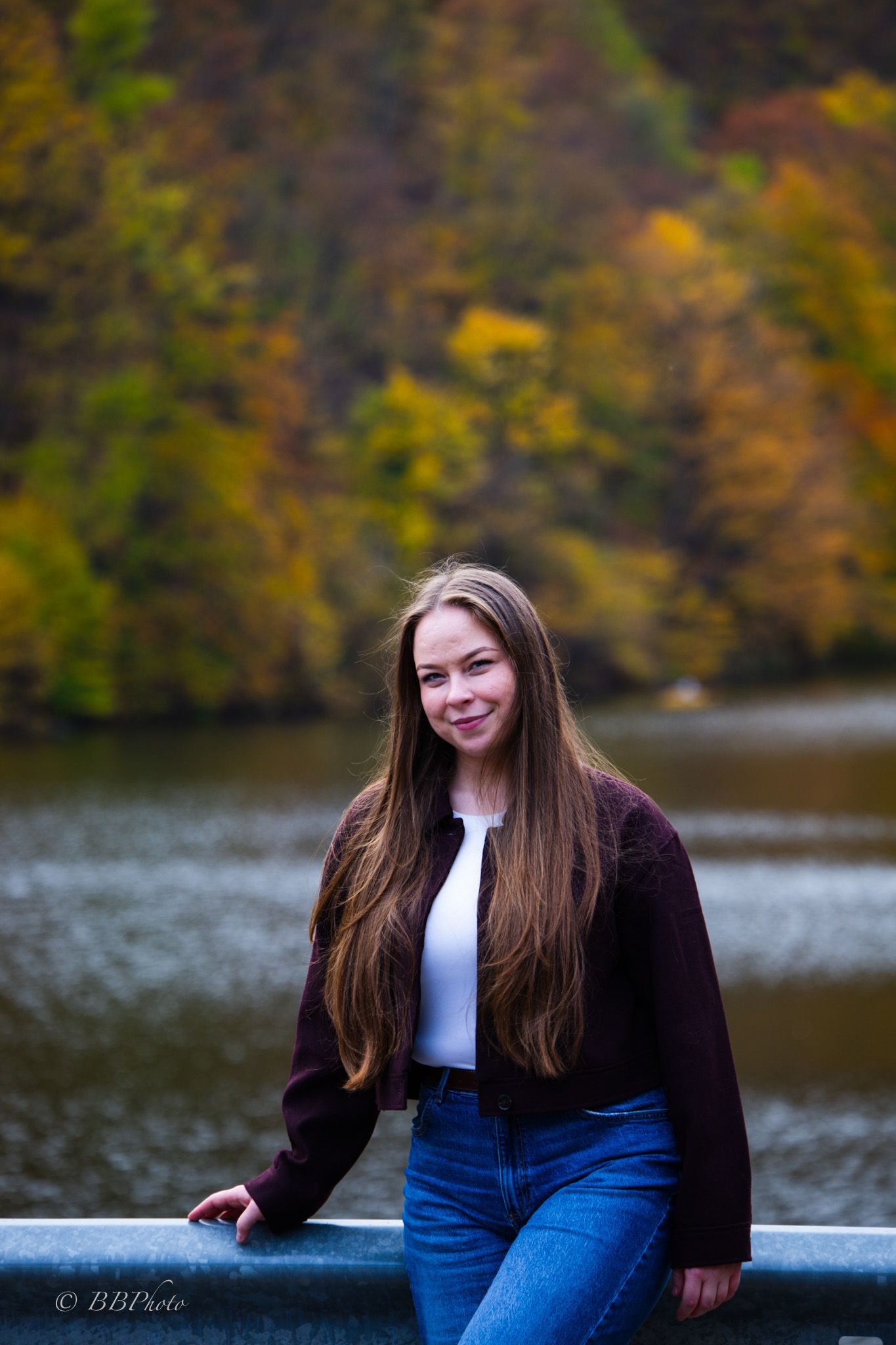 Smiling woman with long brown hair leans on a railing by a lake with colorful autumn trees in the background.