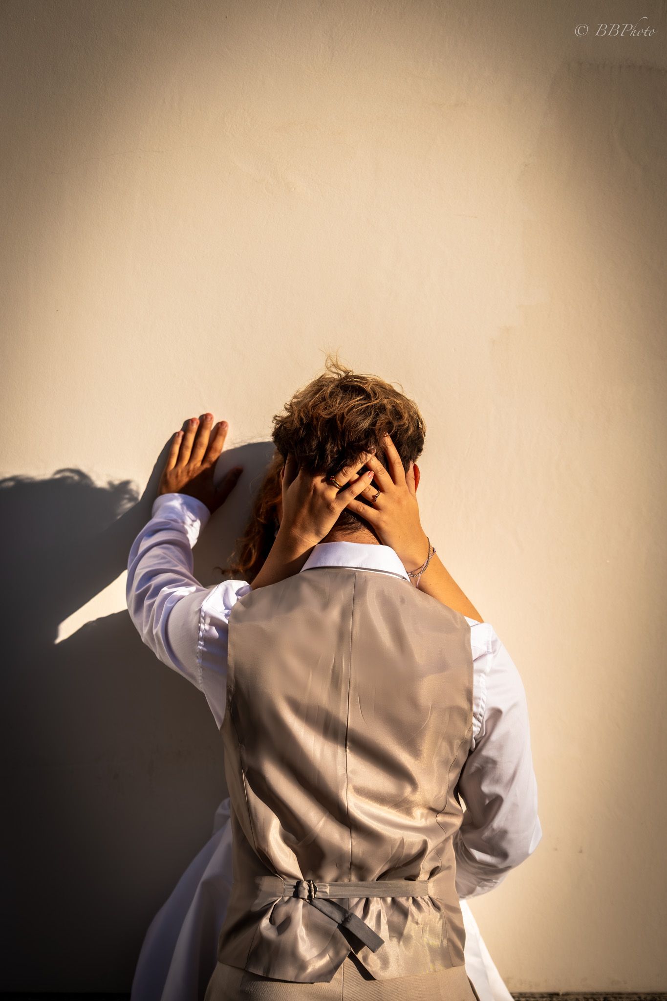 A couple sharing a romantic kiss against a sunlit wall, the groom in a grey vest with his back to the camera.