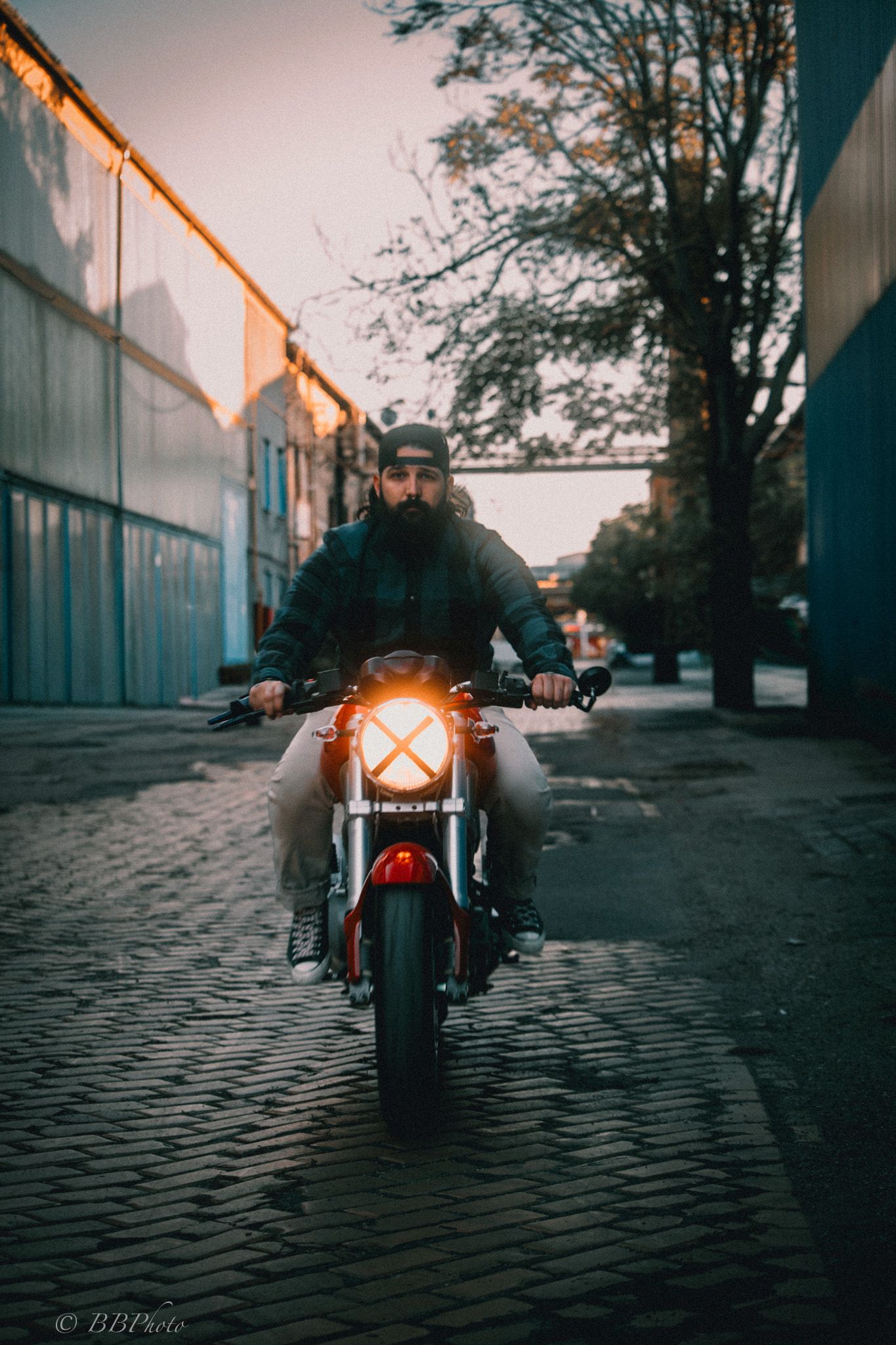 Bearded man in a cap rides a red motorcycle with a glowing headlight down a cobblestone street between industrial buildings at dusk.