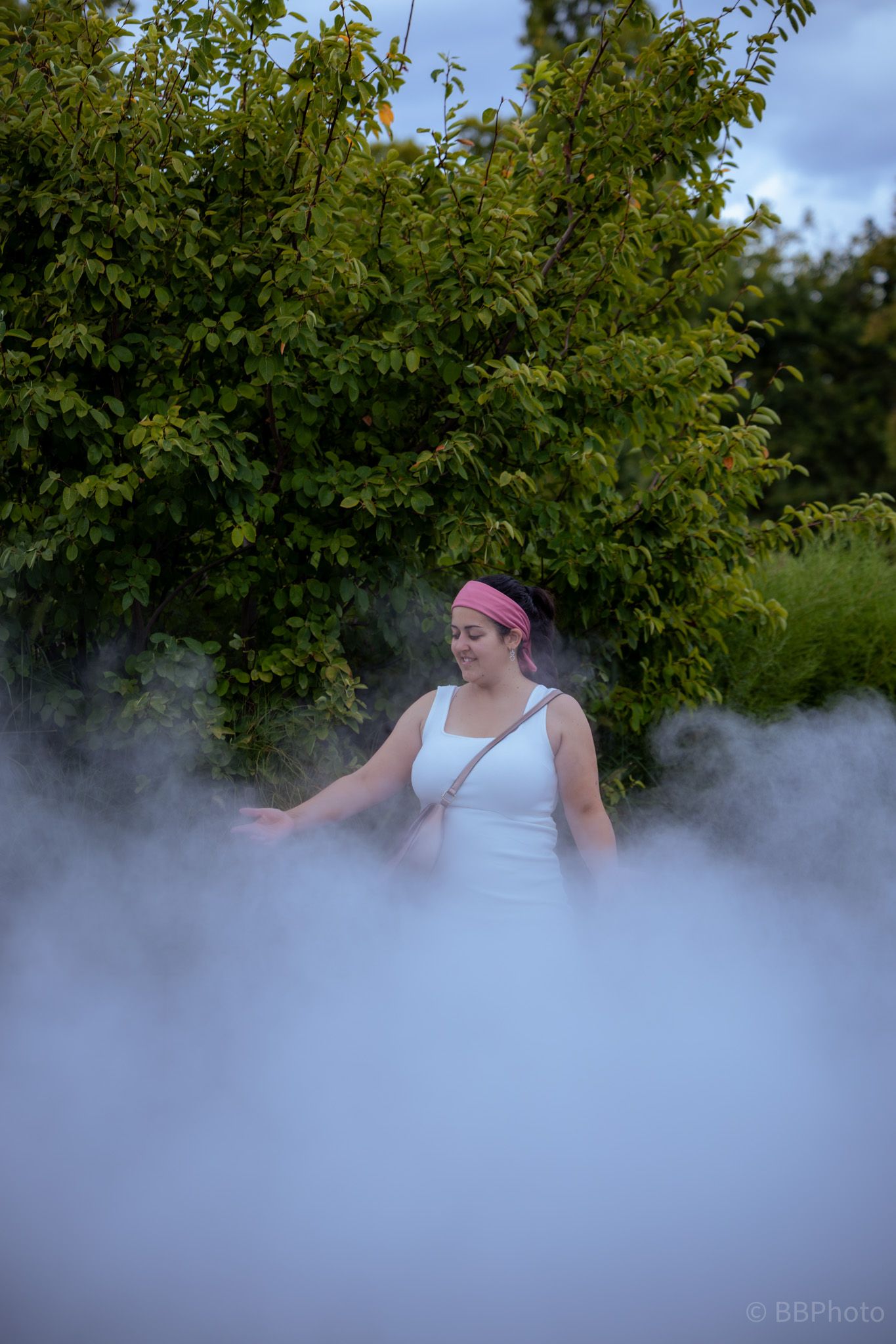 Woman in a white dress standing surrounded by thick white smoke or fog in a lush green outdoor setting.