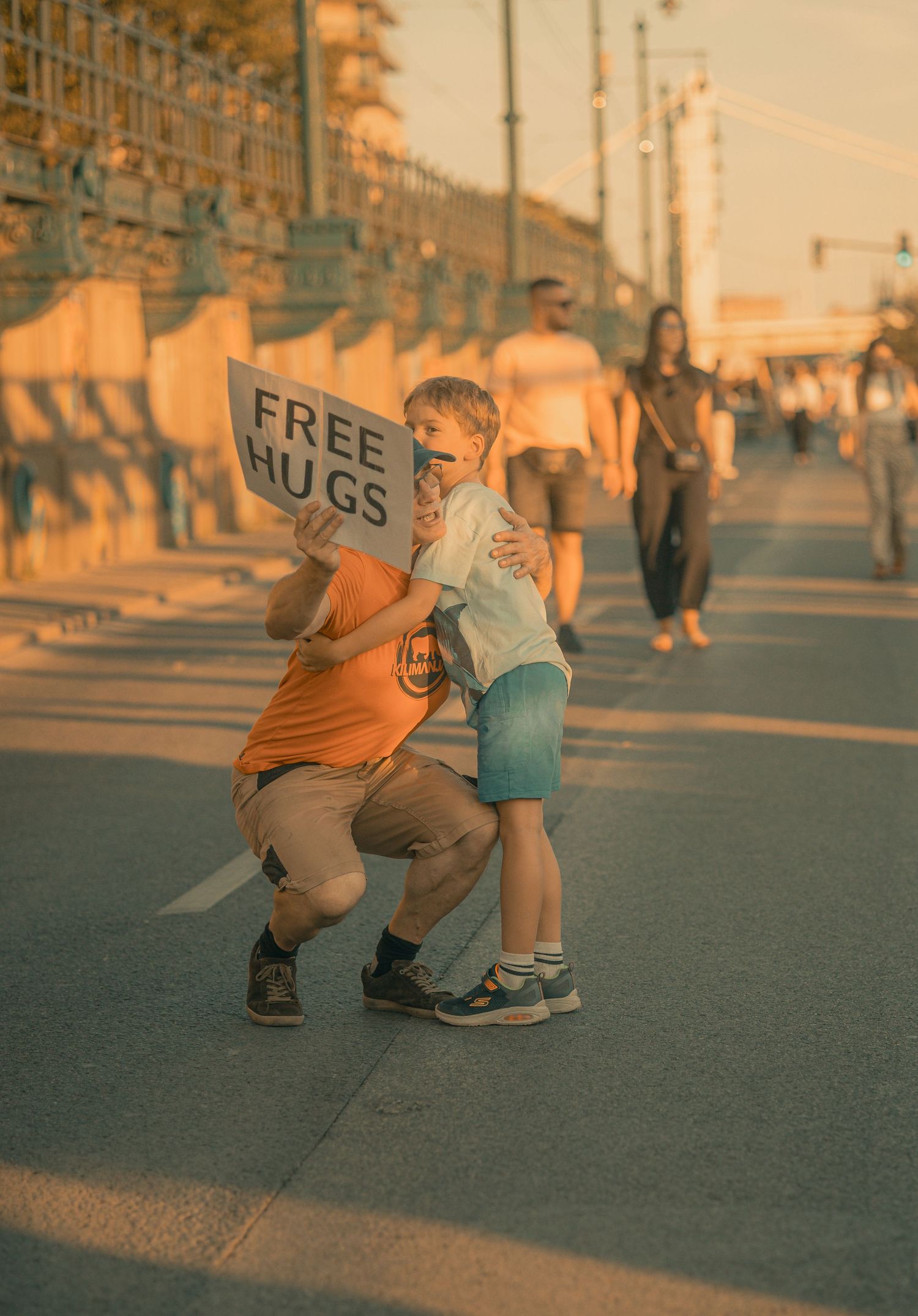 A child holds a Free Hugs sign while hugging another kid on a sunny boardwalk with people walking in the background.