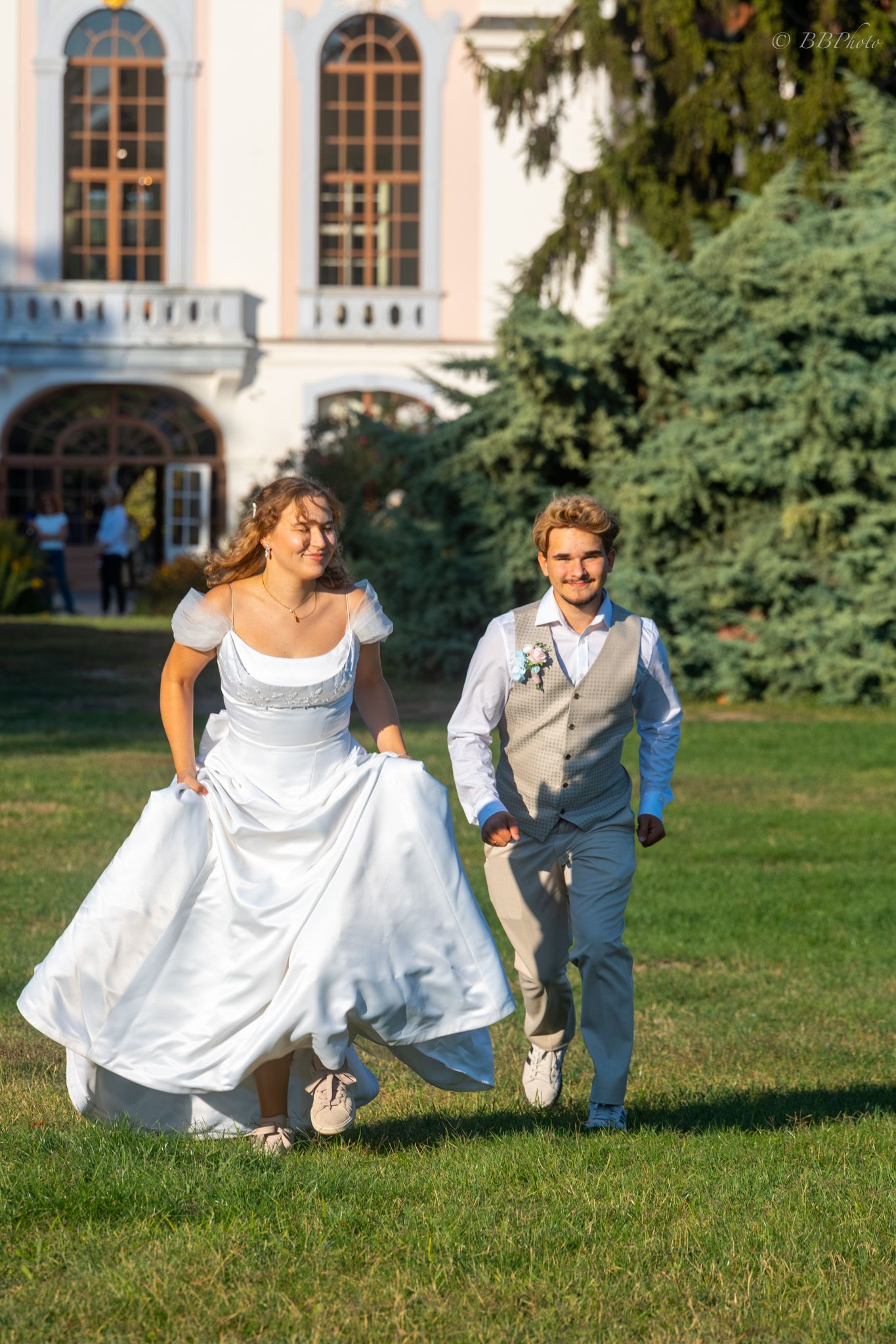 Joyful bride in white gown and groom in gray vest running hand-in-hand on green lawn outside elegant estate.
