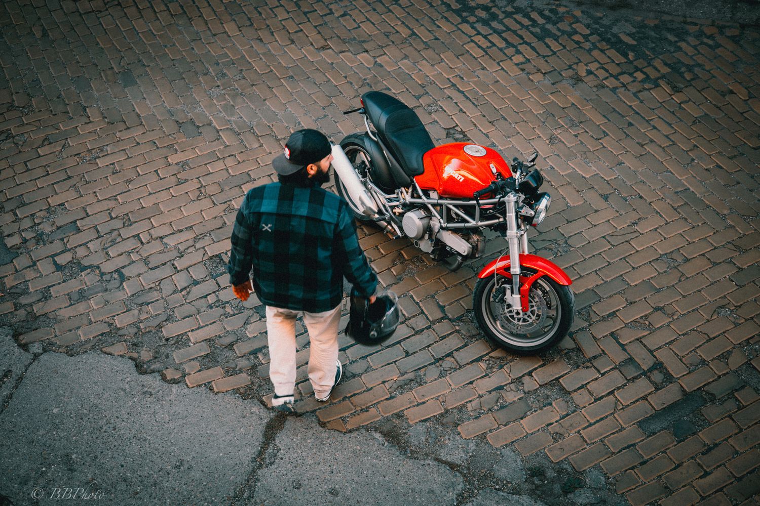 Man in a green plaid jacket and cap standing on a brick street beside a red Ducati Monster motorcycle, holding a helmet.