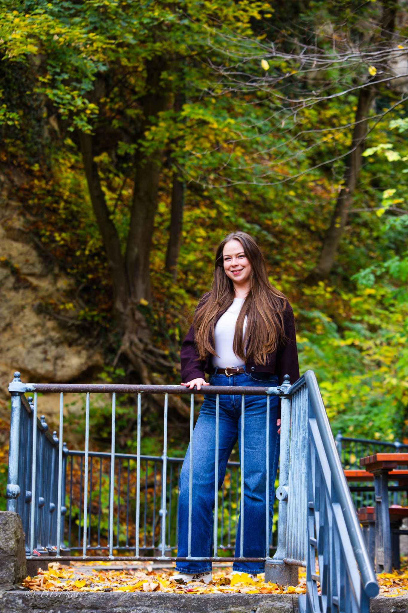 Smiling woman with long brown hair in jeans and a dark cardigan stands on a metal railing staircase surrounded by colorful autumn foliage.