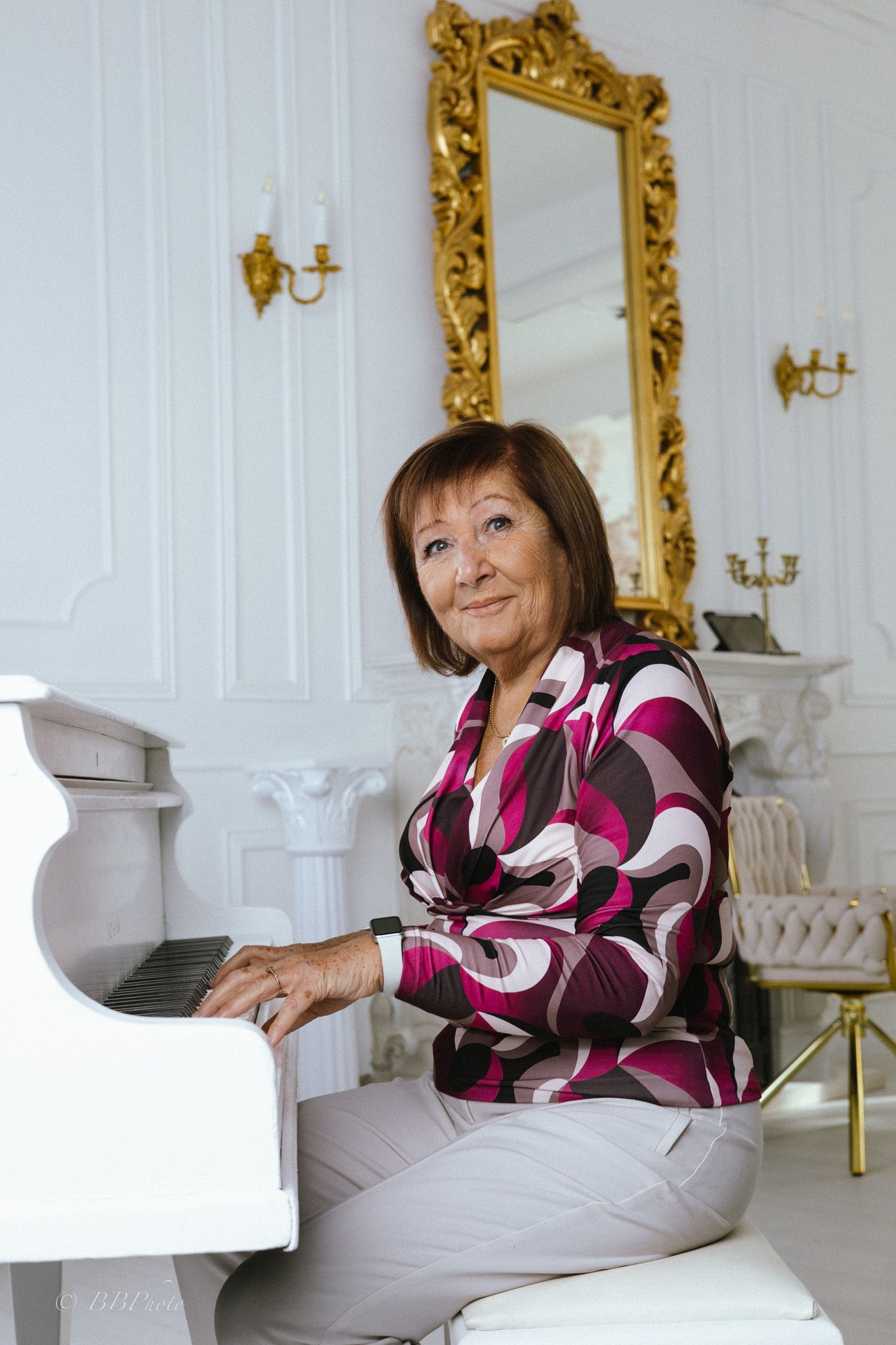 Smiling older woman in a patterned magenta blouse sits at a white piano in an elegant room with a gilded mirror and wall sconces.