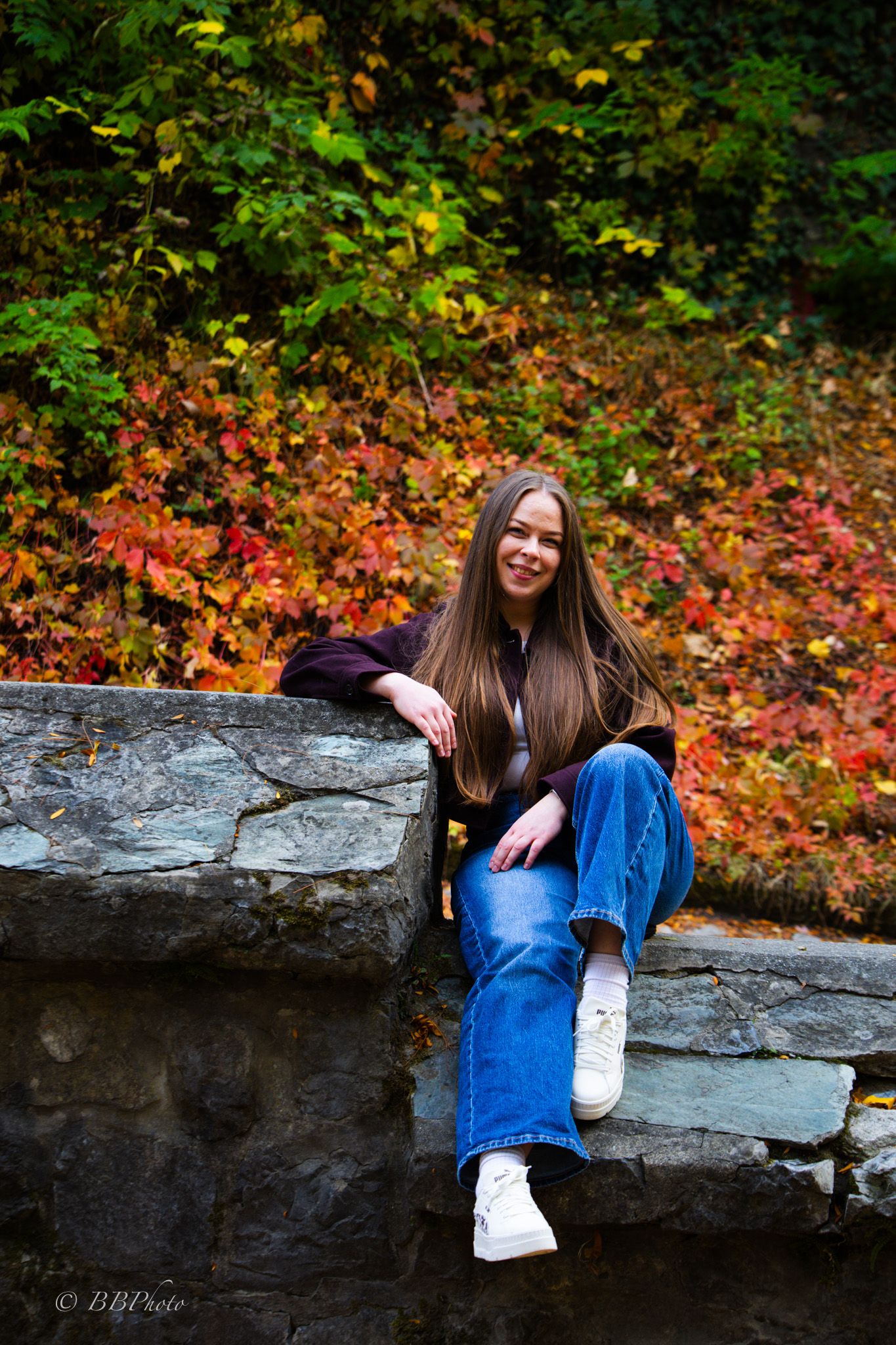Smiling young woman with long brown hair sits on a stone wall in blue jeans and white sneakers, with colorful red autumn foliage behind.