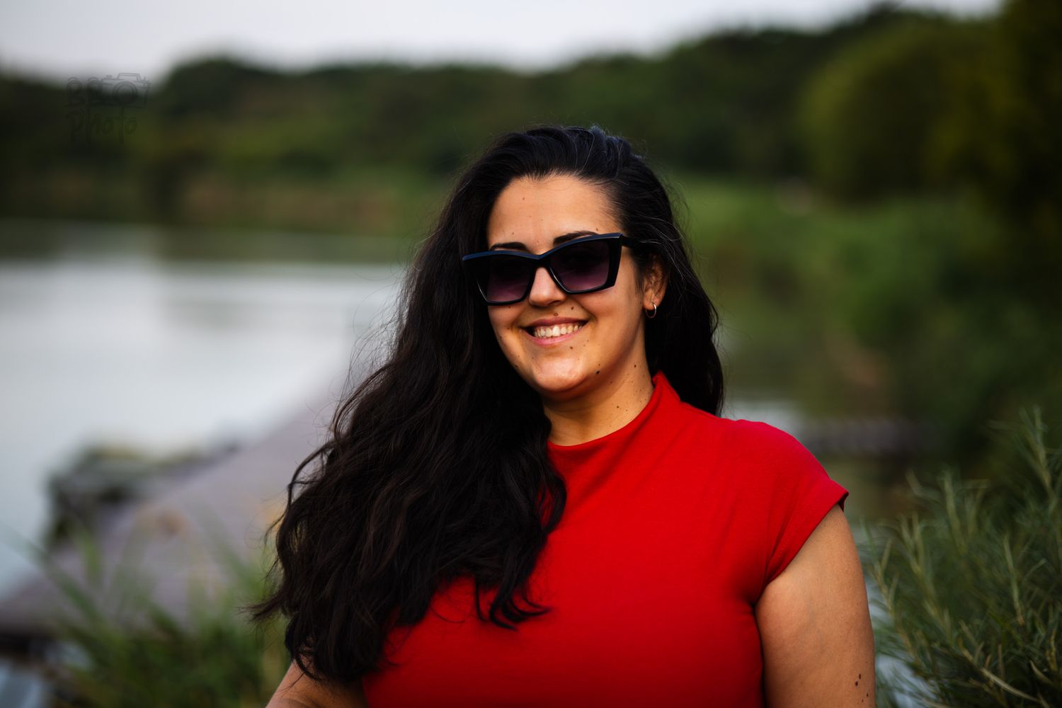 Smiling woman with long dark hair and sunglasses wearing a red top near a lake.