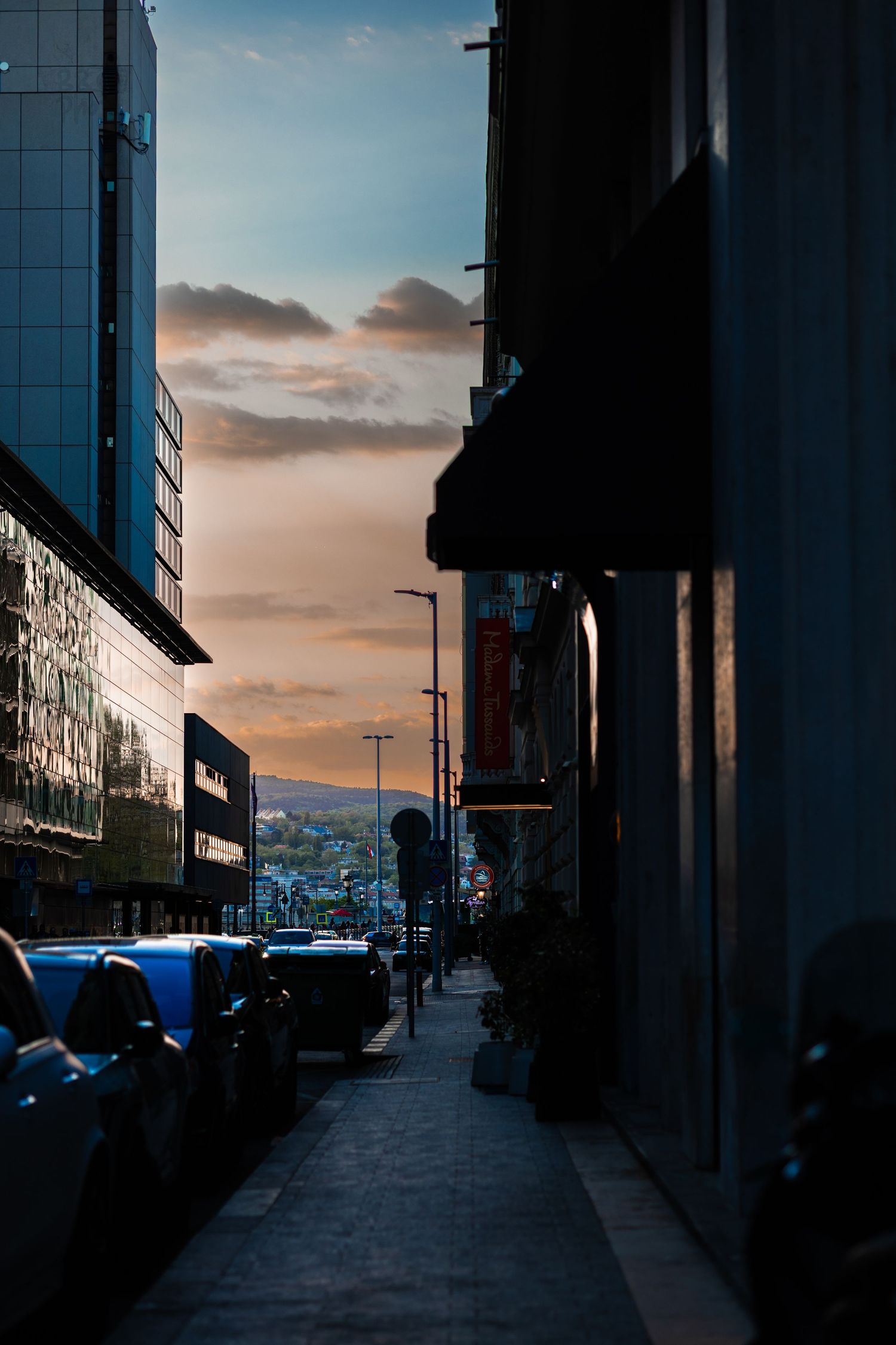 A narrow urban alley at dusk with parked cars, city buildings, and a colorful sunset sky with clouds in the background.