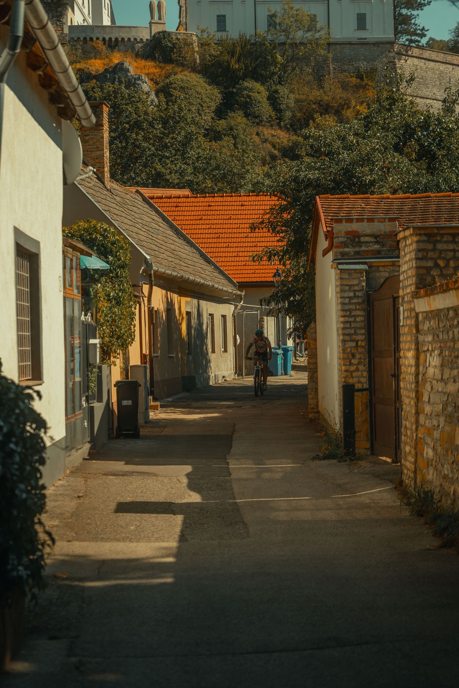 Narrow sunlit alley with historic low-rise buildings, red-tiled roofs, and a hilltop church in a European village.