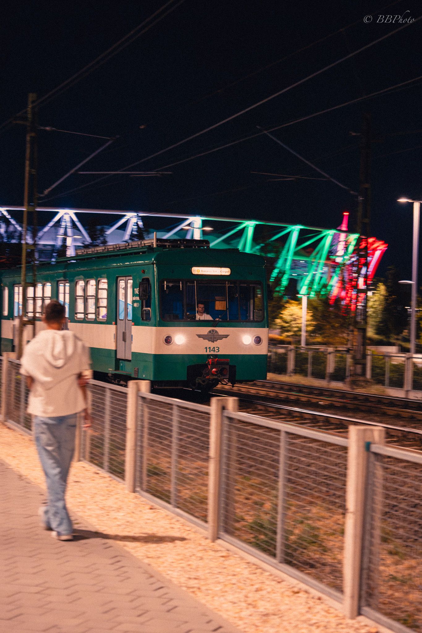 Green tram arriving at night station with colorful stadium lights in background, pedestrian walking along platform.