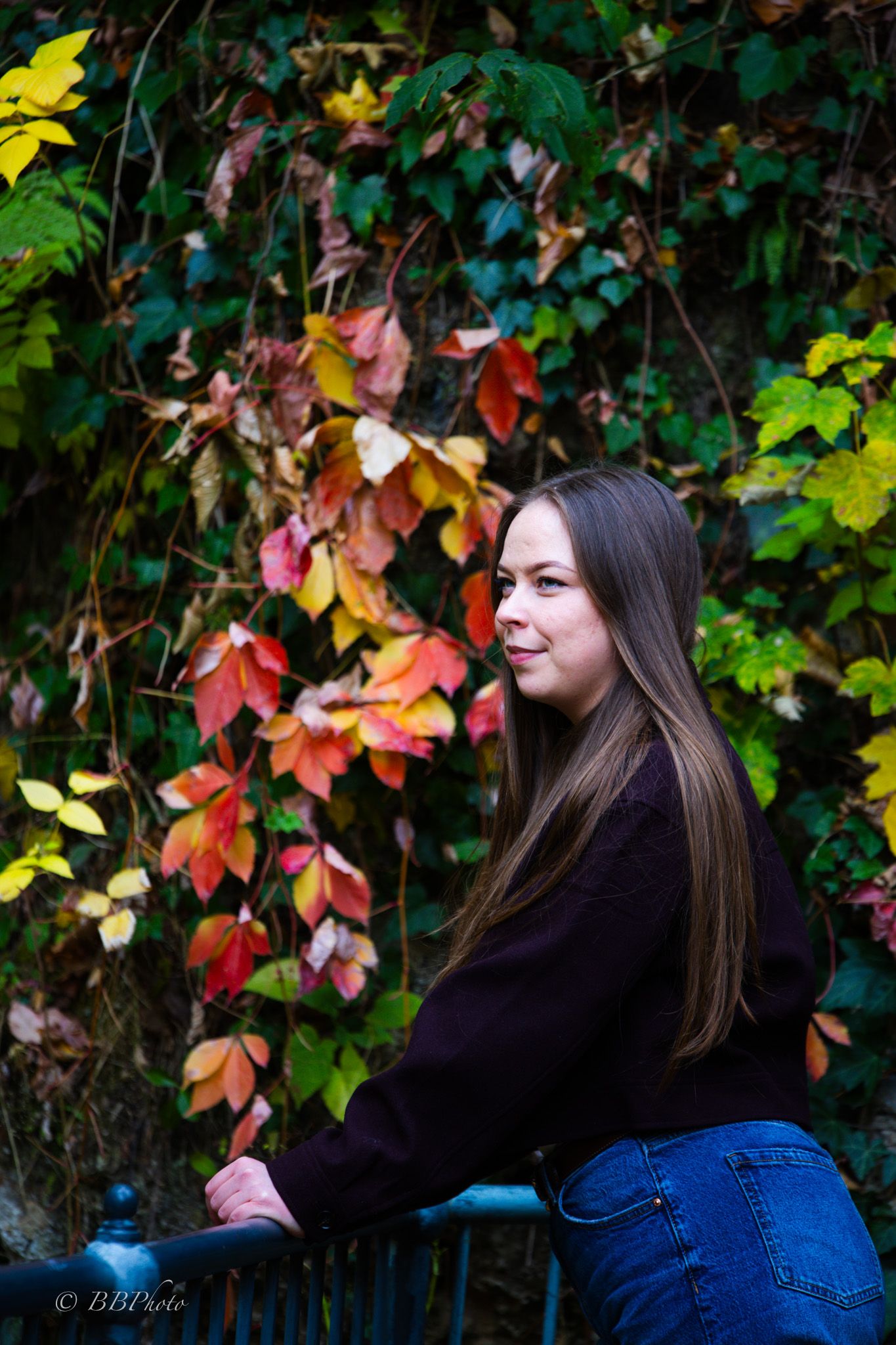 Young woman with long brown hair leans on a blue railing beside a wall of red and orange autumn ivy leaves.