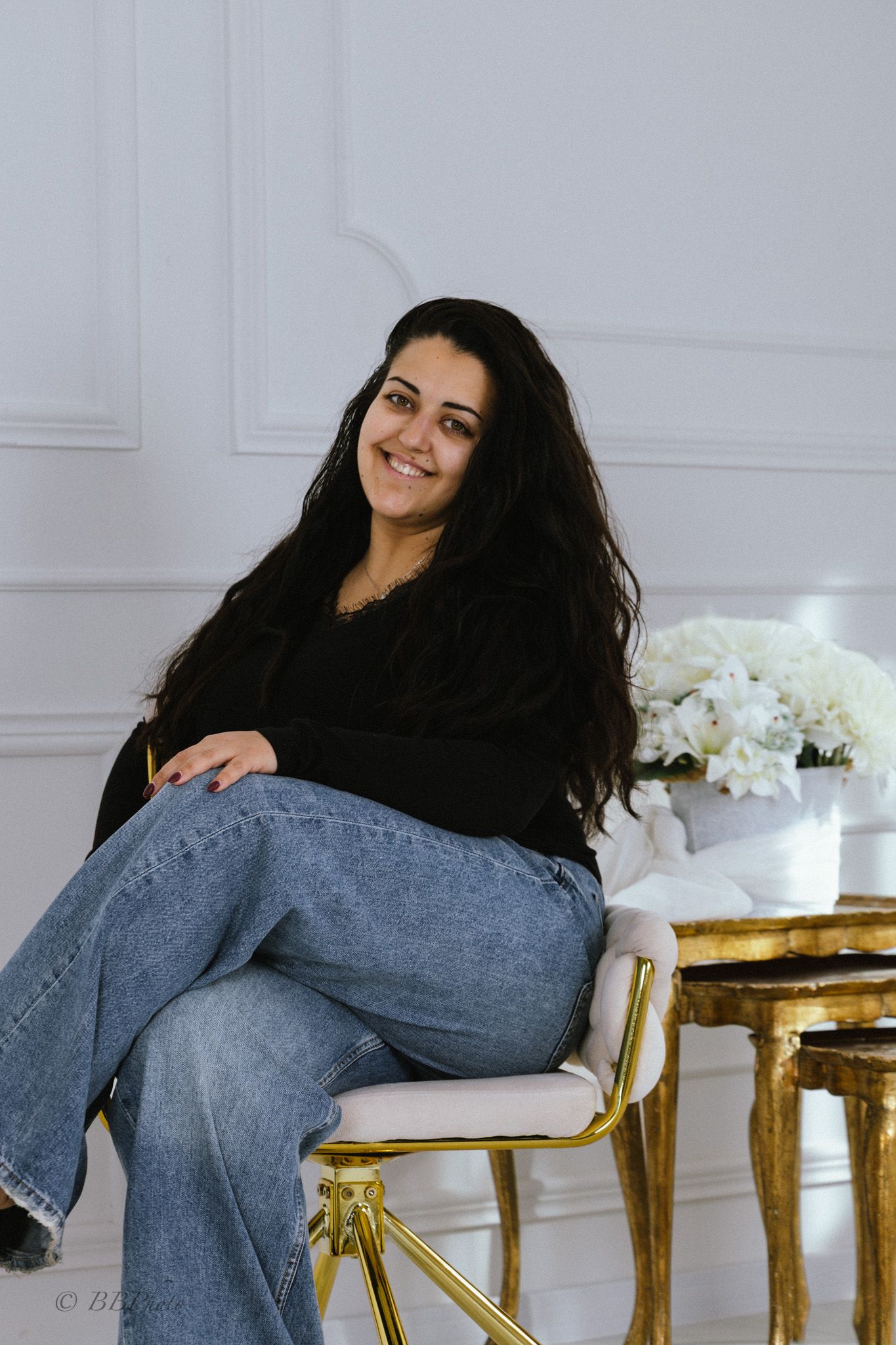 Smiling woman with long dark wavy hair sits on a gold and cream chair in a bright room with white paneled walls and white flowers.