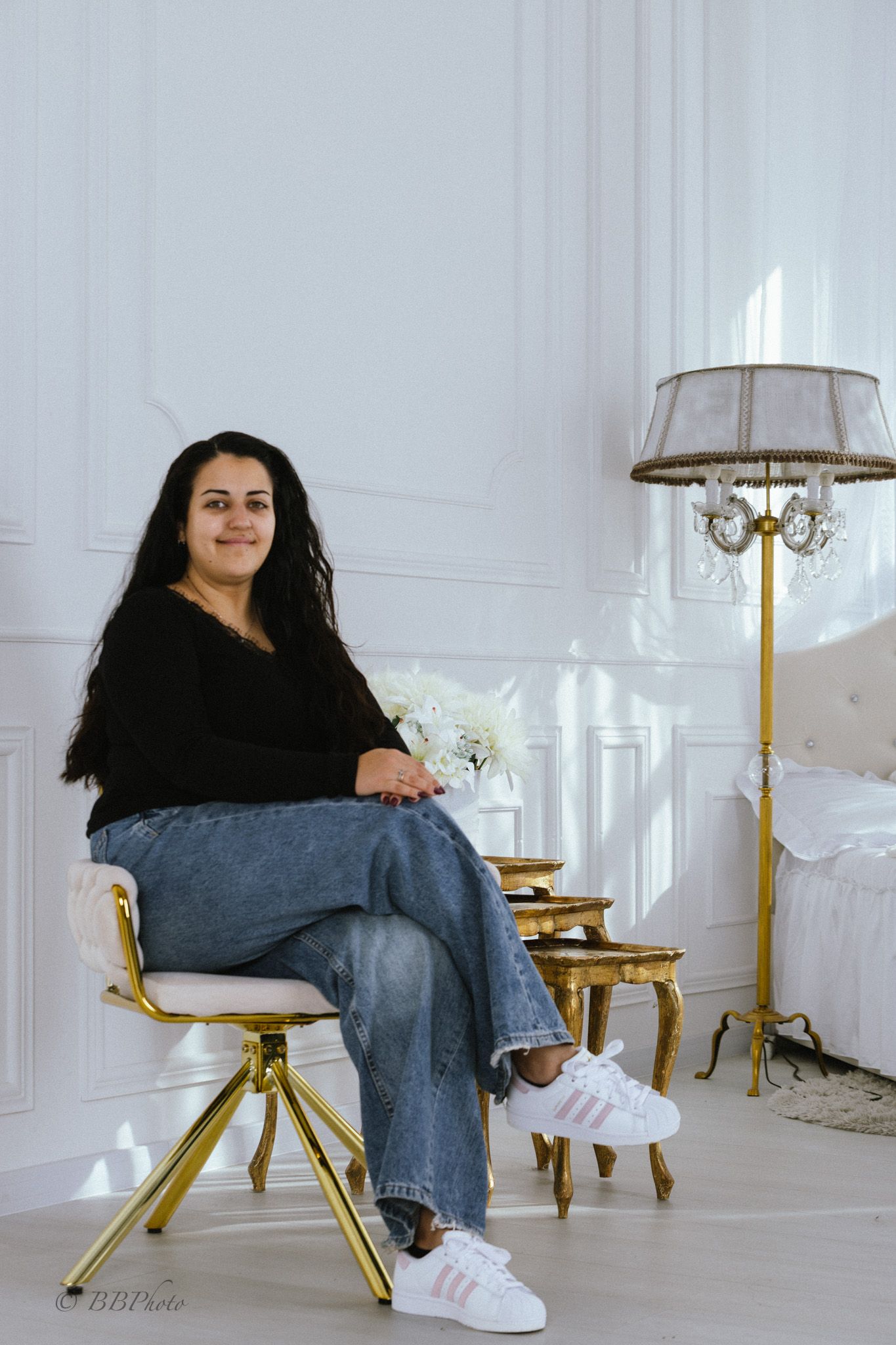 Smiling woman with long dark hair sits on a gold modern chair in a bright white room with a chandelier floor lamp and gold side tables.