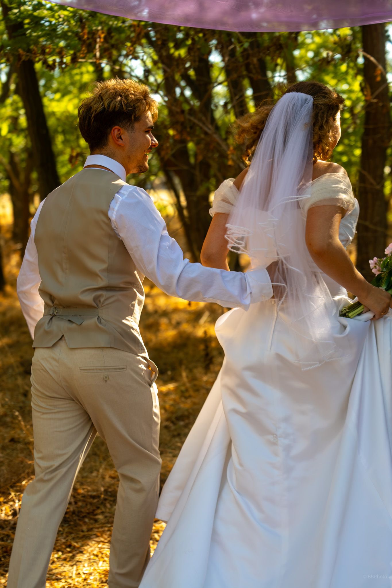 A groom in a beige suit walks with his bride in a white gown and veil through a sunlit forest setting.