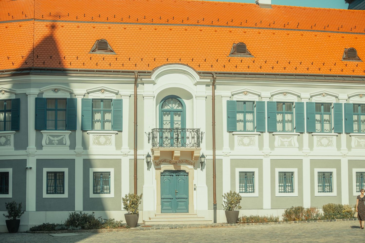 Elegant Baroque palace facade with teal shutters, ornate balcony, and orange roof in a European town square.
