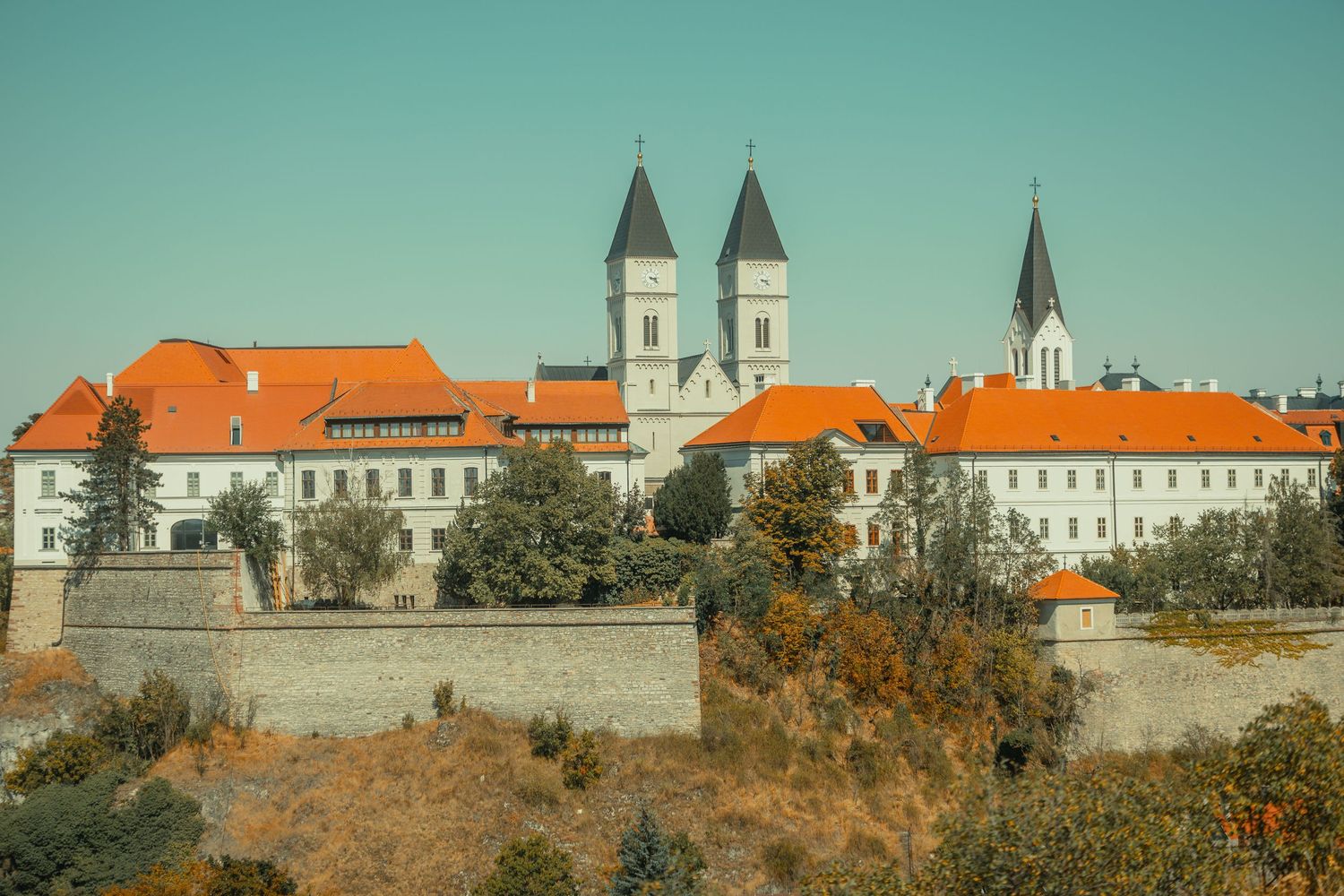 Historic Veszprem Castle district with white buildings, orange rooftops, and medieval church towers against clear sky.