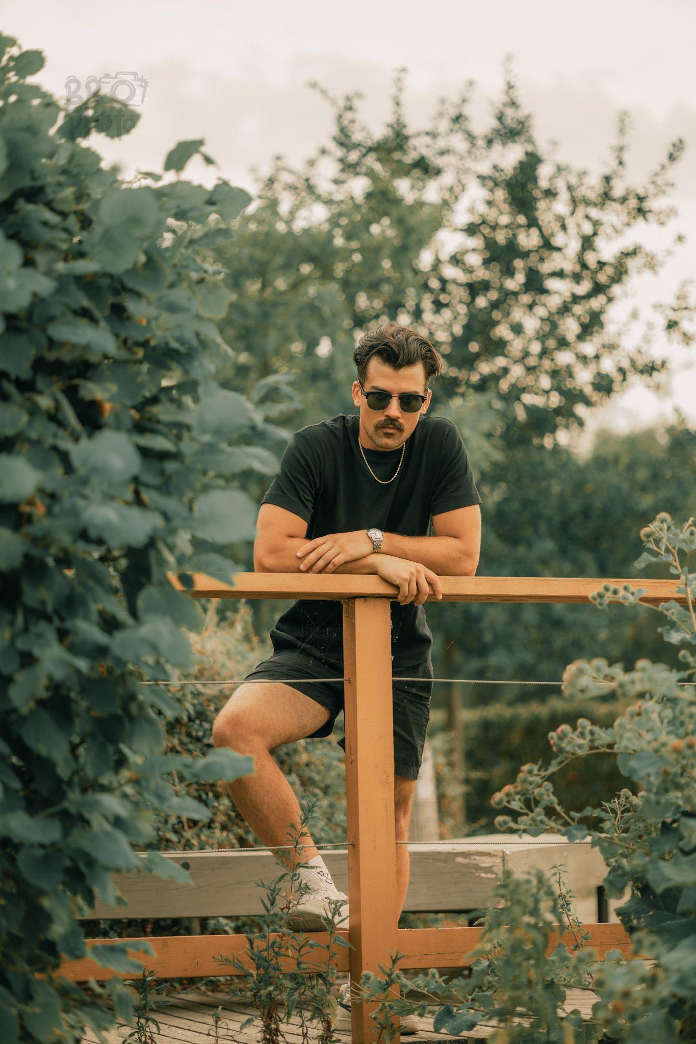 Man with mustache and sunglasses leaning on a wooden railing surrounded by green foliage.
