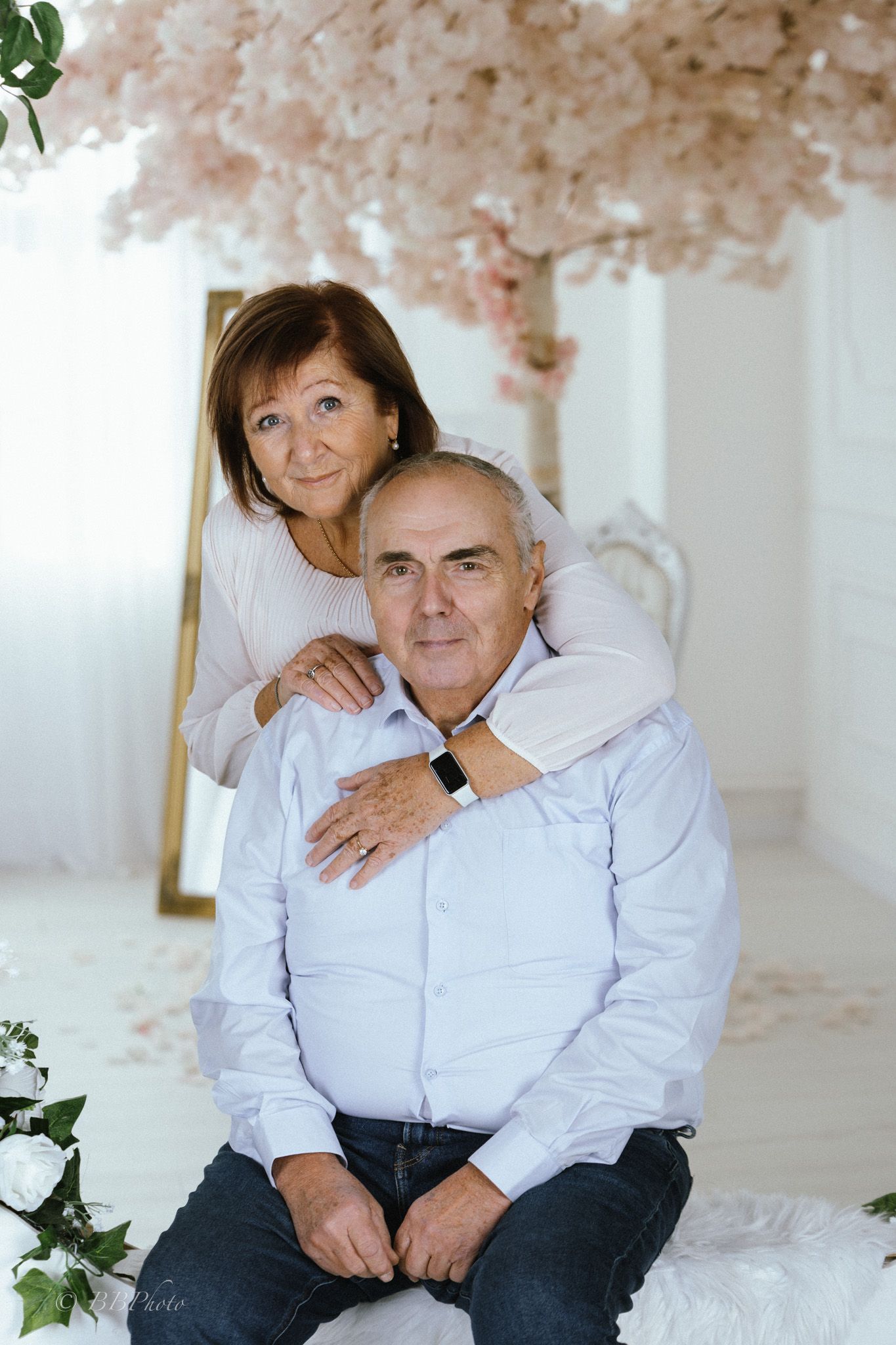 Smiling older couple posing for a portrait in a bright studio with pink blossom tree decor, white florals, and a gold-framed mirror.