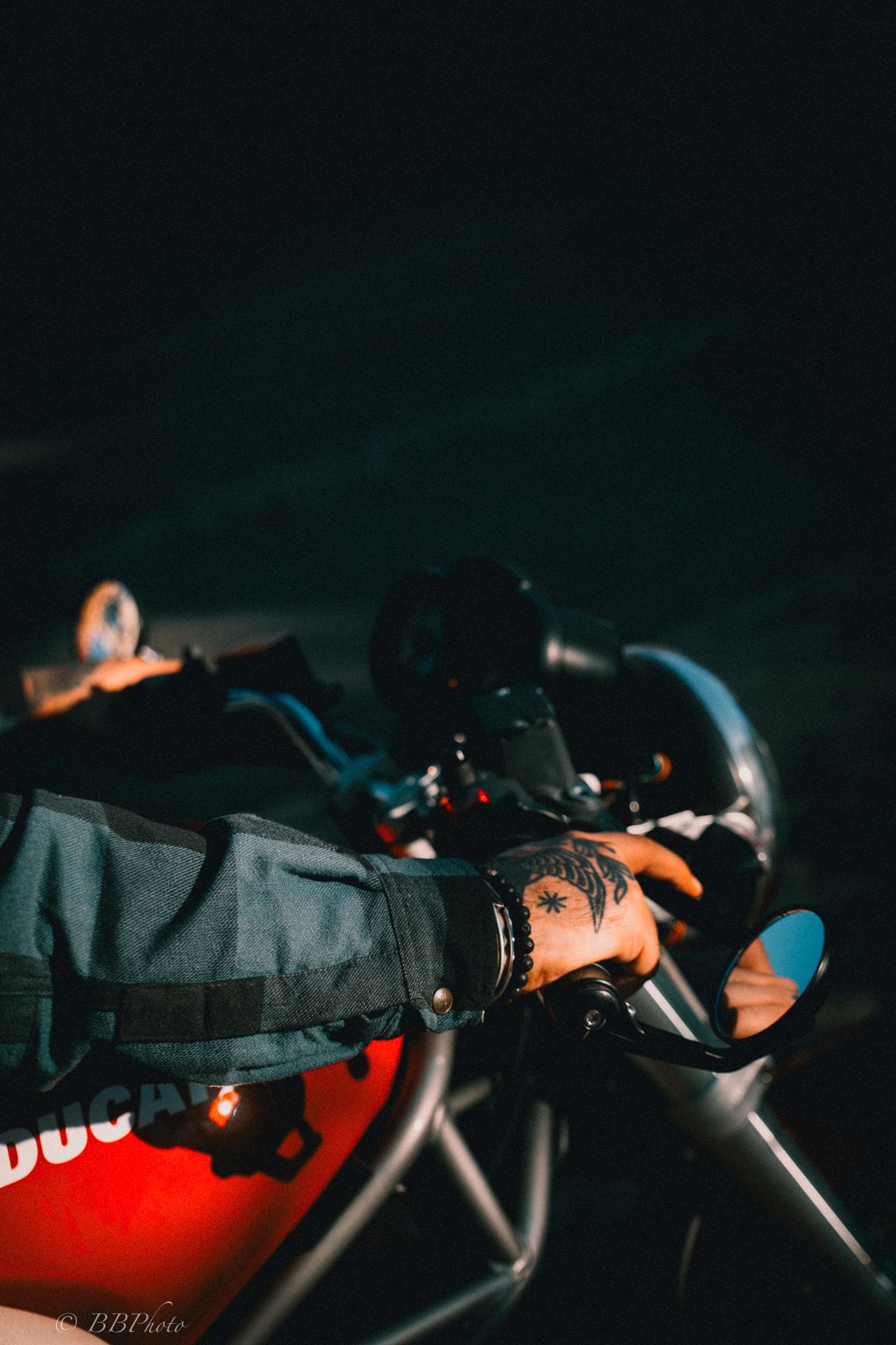 Tattooed hand gripping the handlebar of a red Ducati motorcycle at night, with a round side mirror and denim jacket sleeve visible.