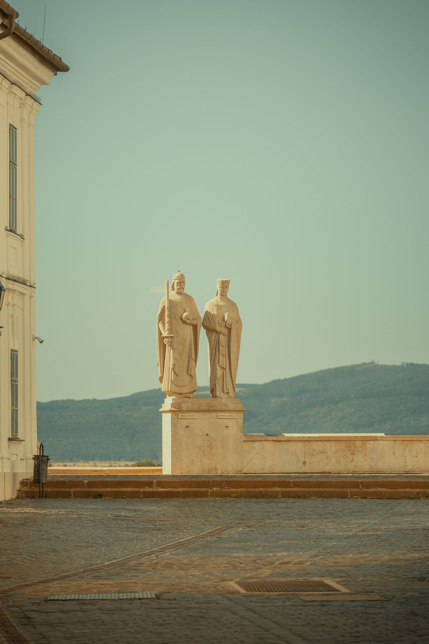Two white stone statues on a pedestal in a sunlit European town square with mountains in the background.