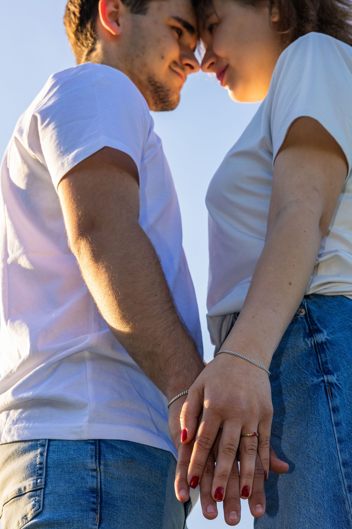 Romantic couple in white t-shirts and jeans holding hands, touching foreheads against a blue sky.