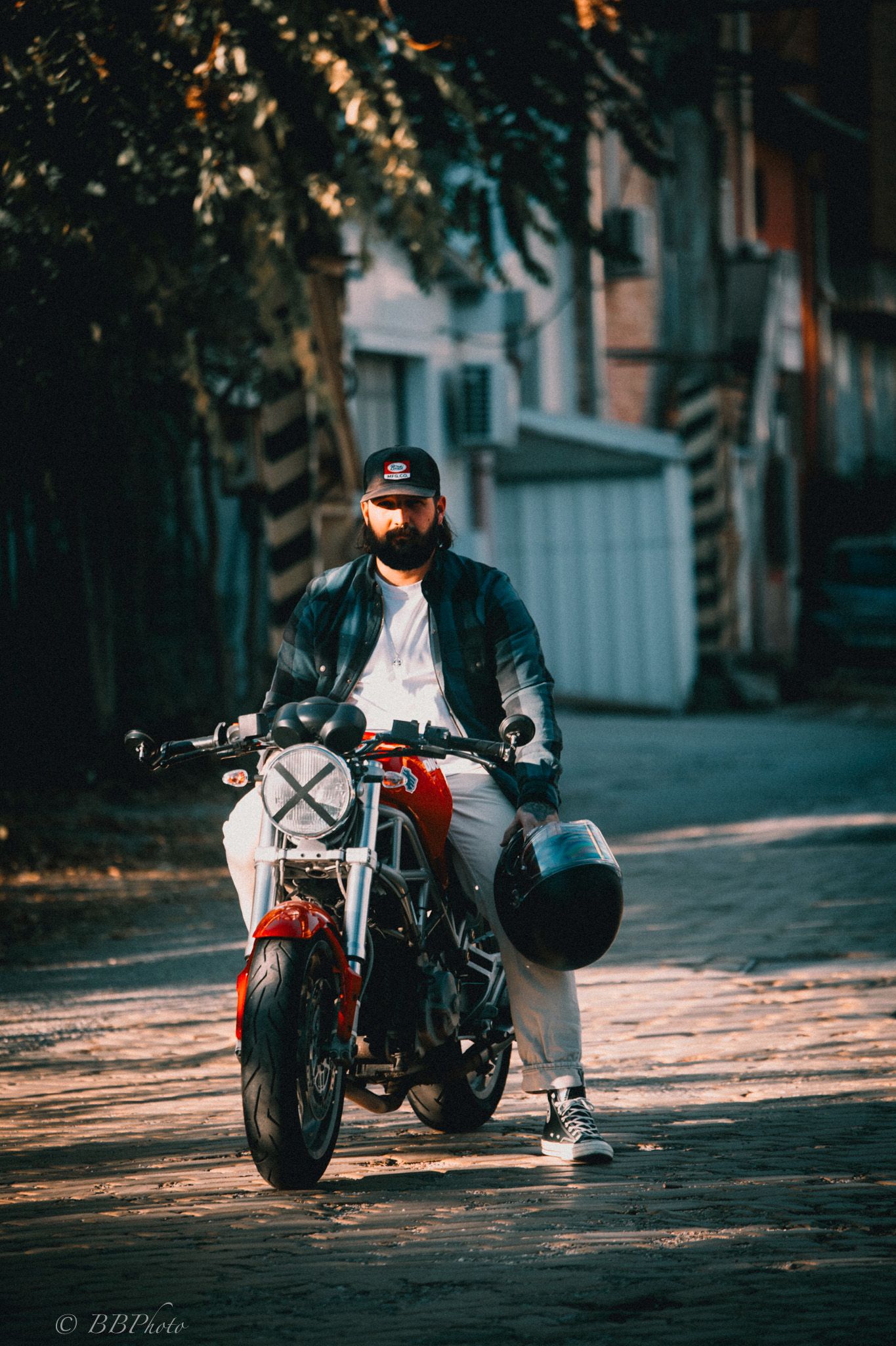 Bearded man in a black cap and leather jacket sits on a red café racer motorcycle holding a helmet on a sunlit cobblestone street.