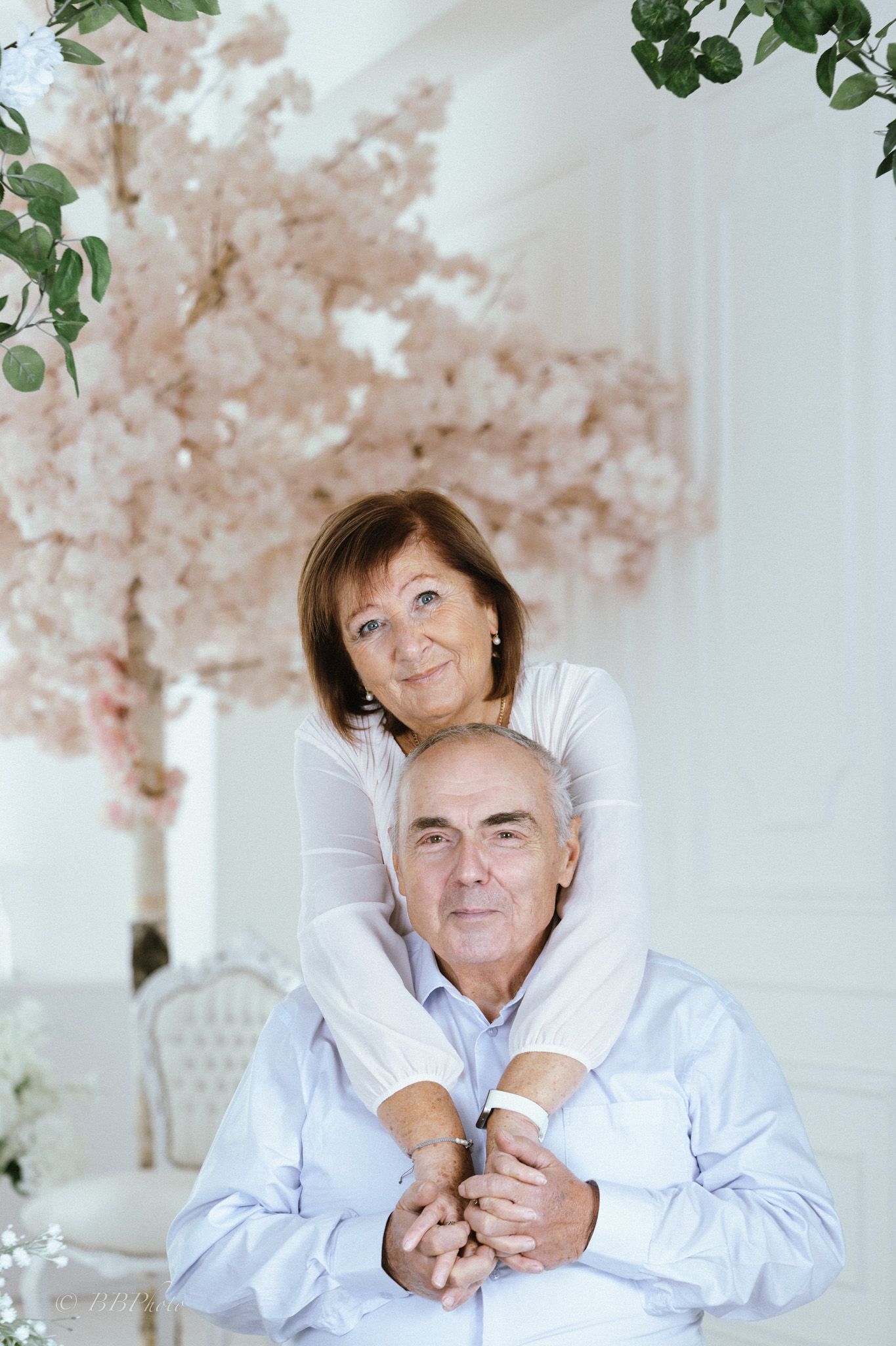 Smiling older woman in a white top embraces a seated older man in a light blue shirt near a blooming pink cherry blossom tree indoors.