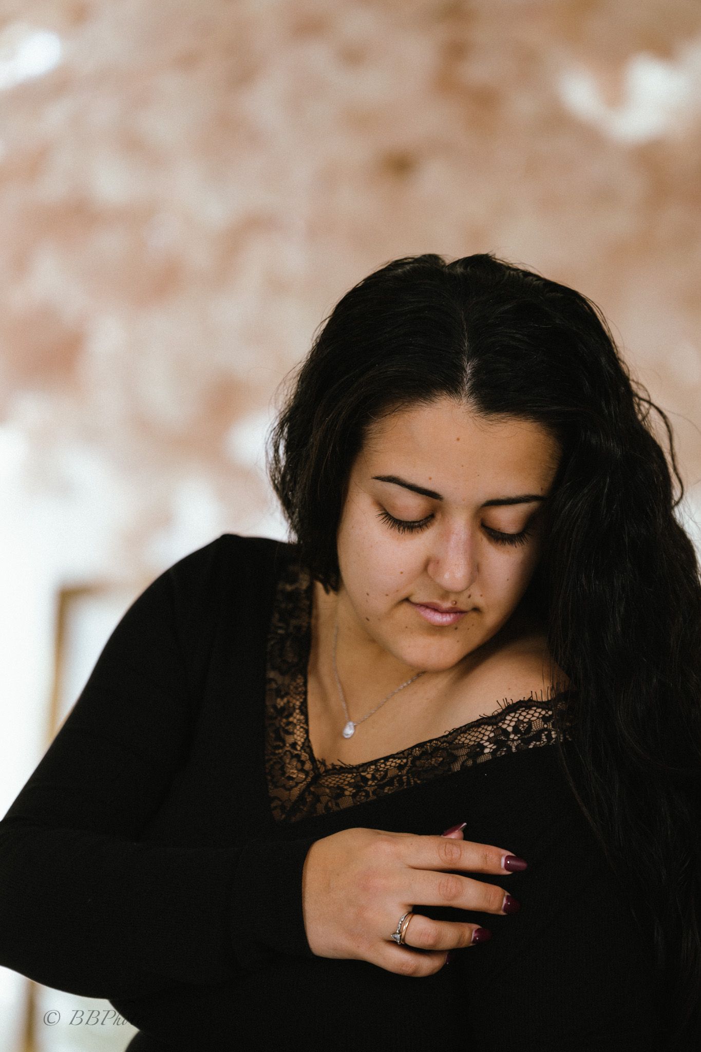 Woman with long dark hair looks down while touching the lace neckline of her black long-sleeve top, wearing a pendant necklace and ring.