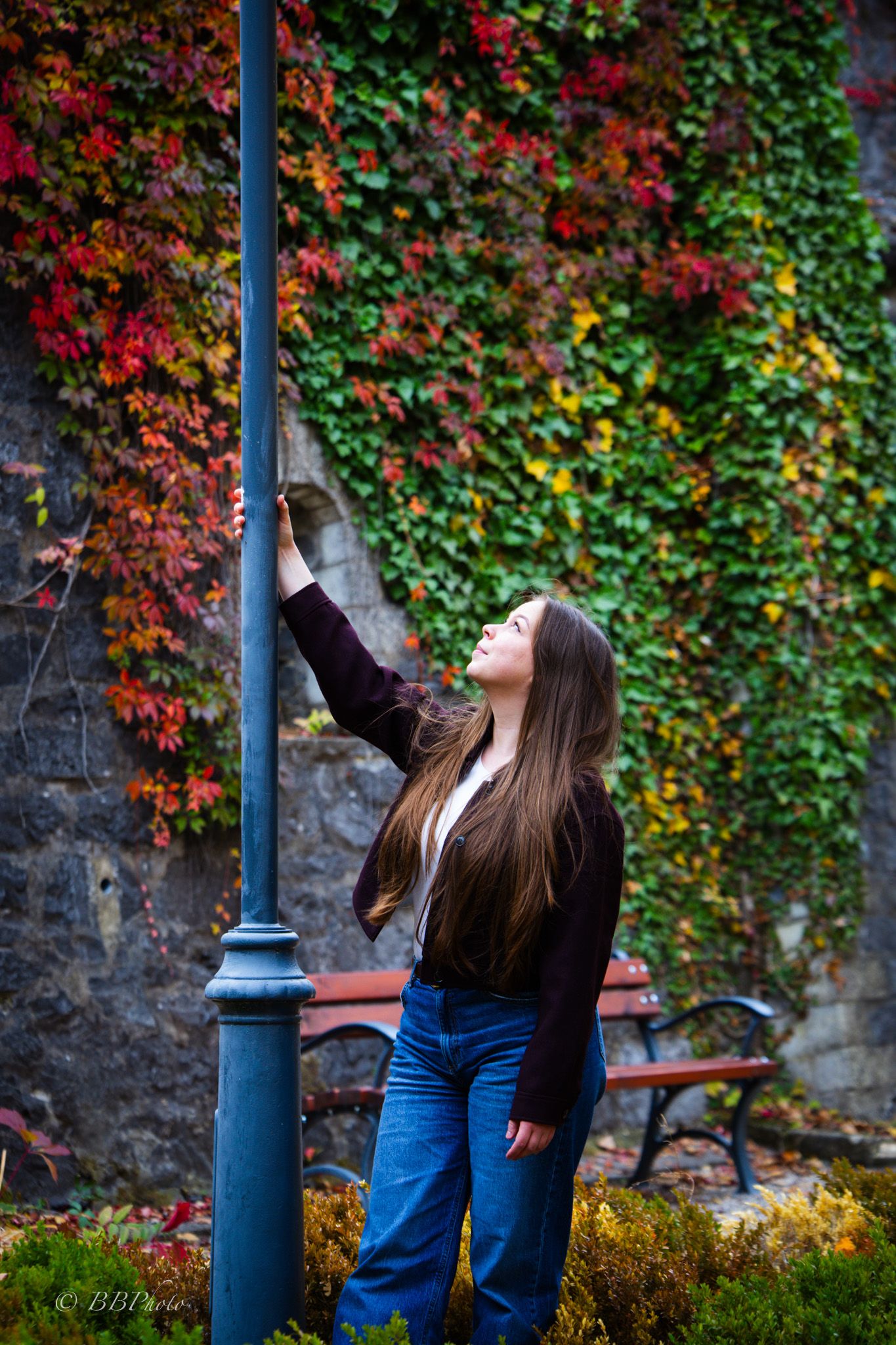 Young woman with long brown hair in jeans and a dark top leans on a lamp post, gazing up beside a wall of red and green autumn ivy.