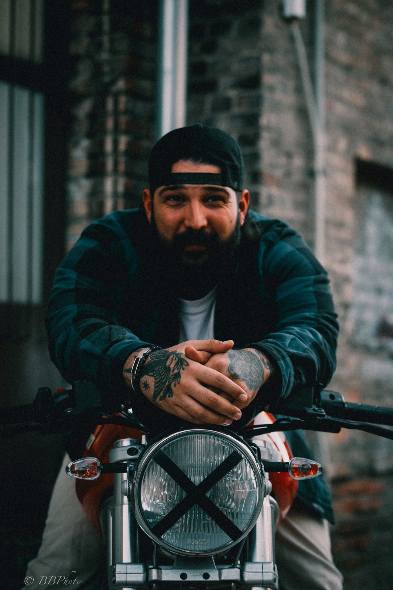 Bearded man in a black cap and plaid jacket leans on the handlebars of a vintage motorcycle with an X-taped headlight near a brick wall.