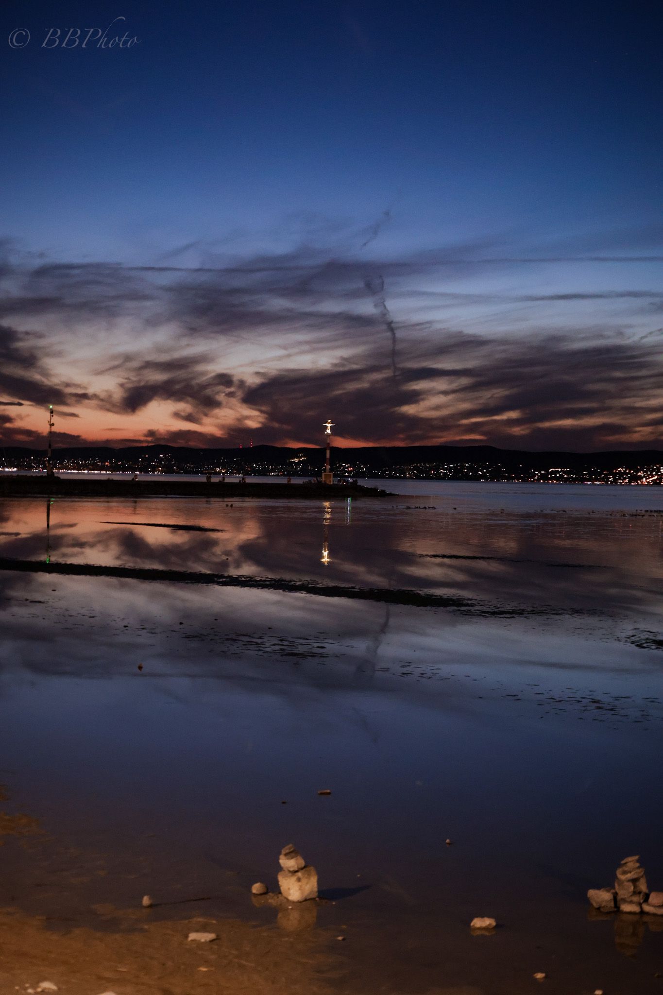 Twilight reflection of a illuminated tower and city lights on calm water at dusk with dramatic clouds.