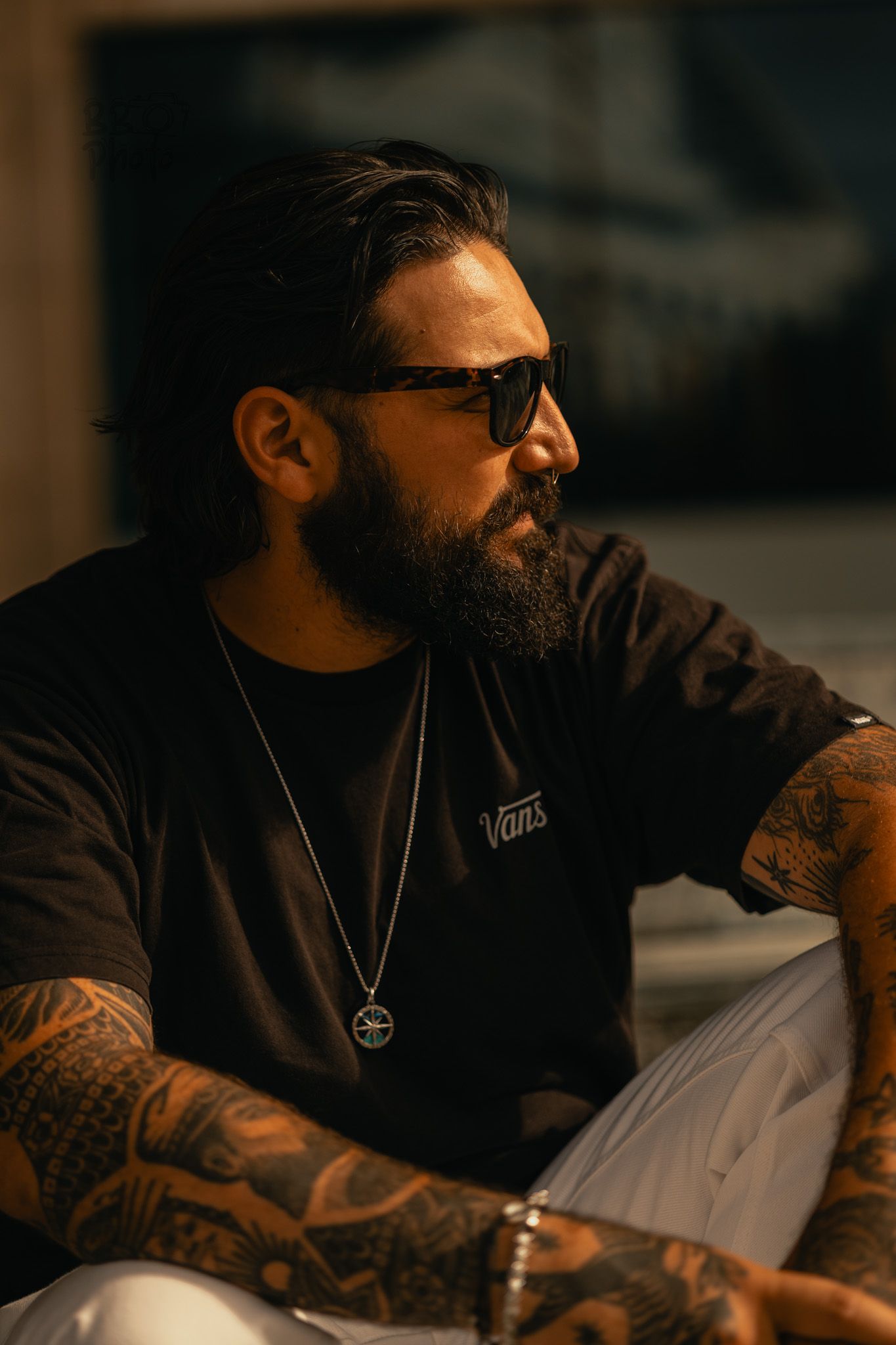 Tattooed bearded man wearing black Vans t-shirt, sunglasses, and silver pendant necklace sitting in warm light.