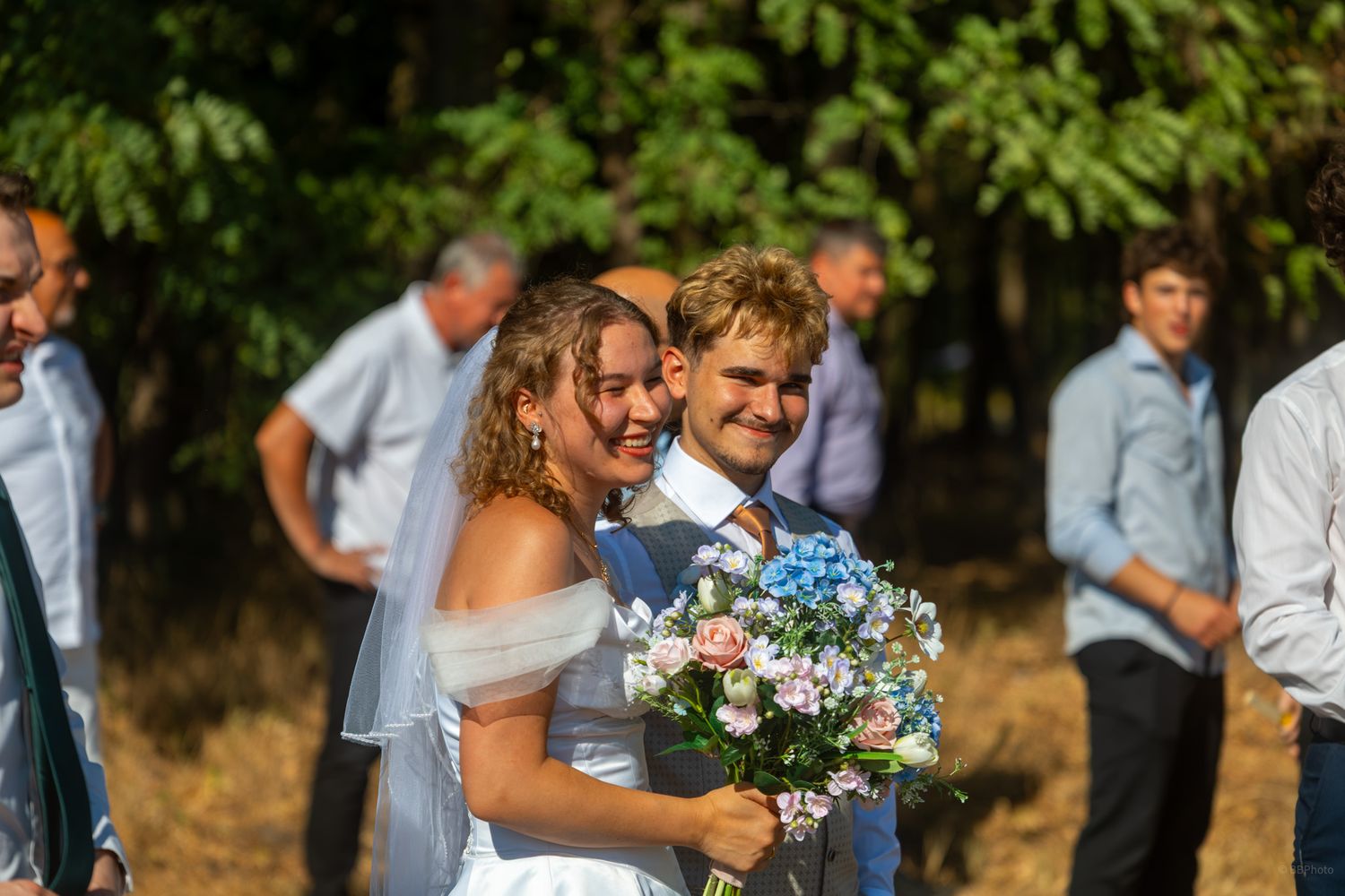 Happy newlyweds smile together outdoors, bride holding pink and blue bouquet, wedding guests in background.