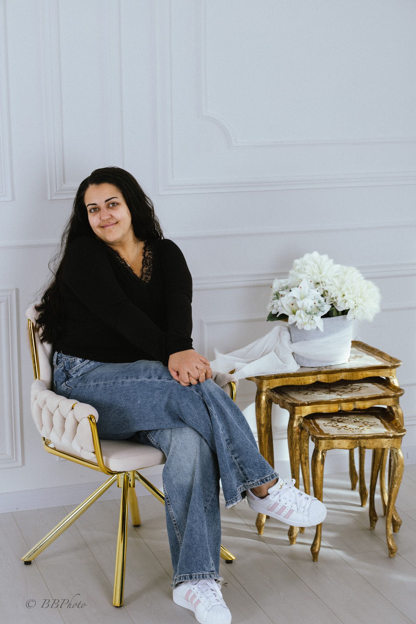 Woman with long dark hair in a black top and jeans sits on a gold-framed chair beside nesting tables with white flowers in a bright studio.