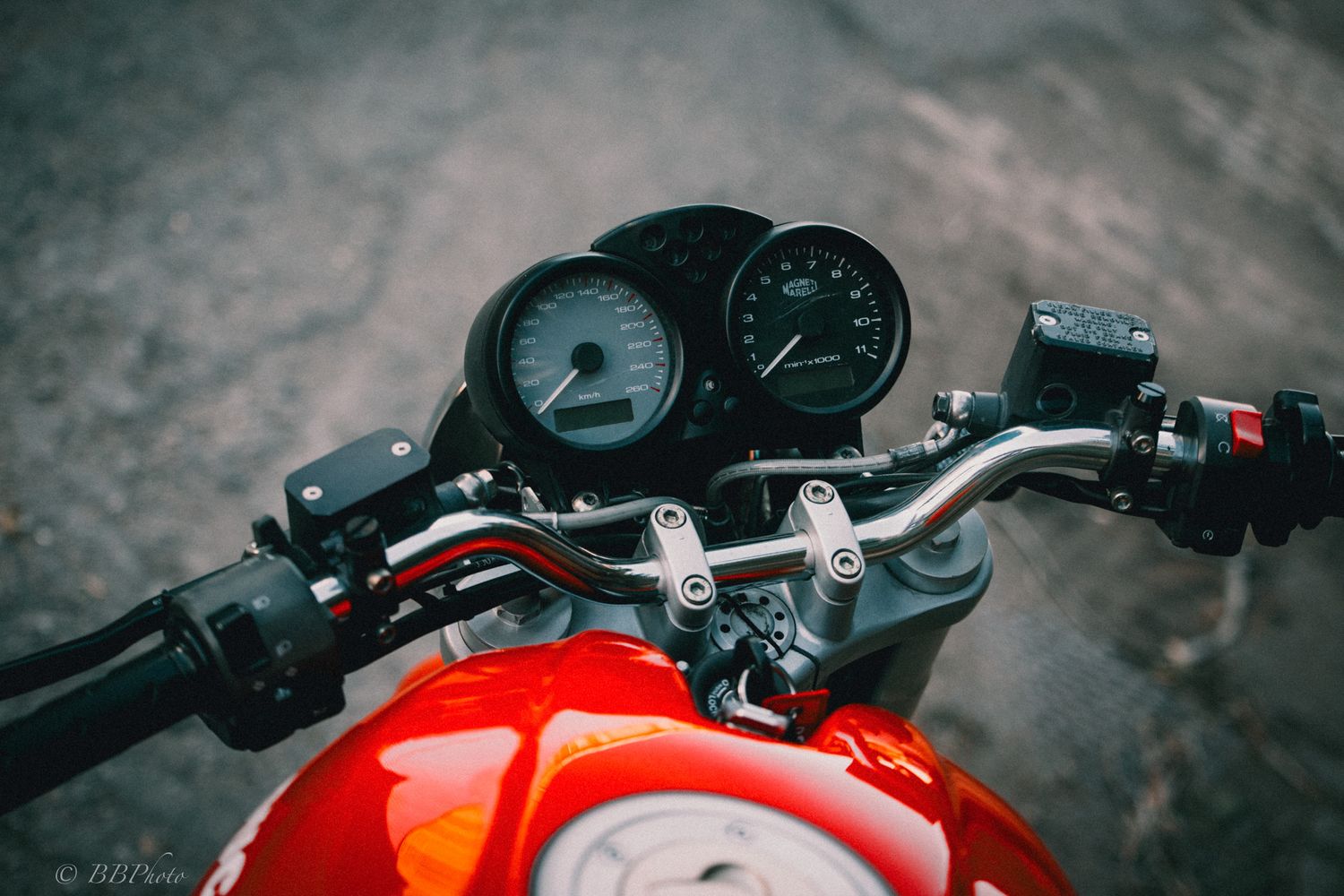 Close-up of a red motorcycle's handlebars, round speedometer and tachometer gauges, and glossy fuel tank on a rough asphalt surface.
