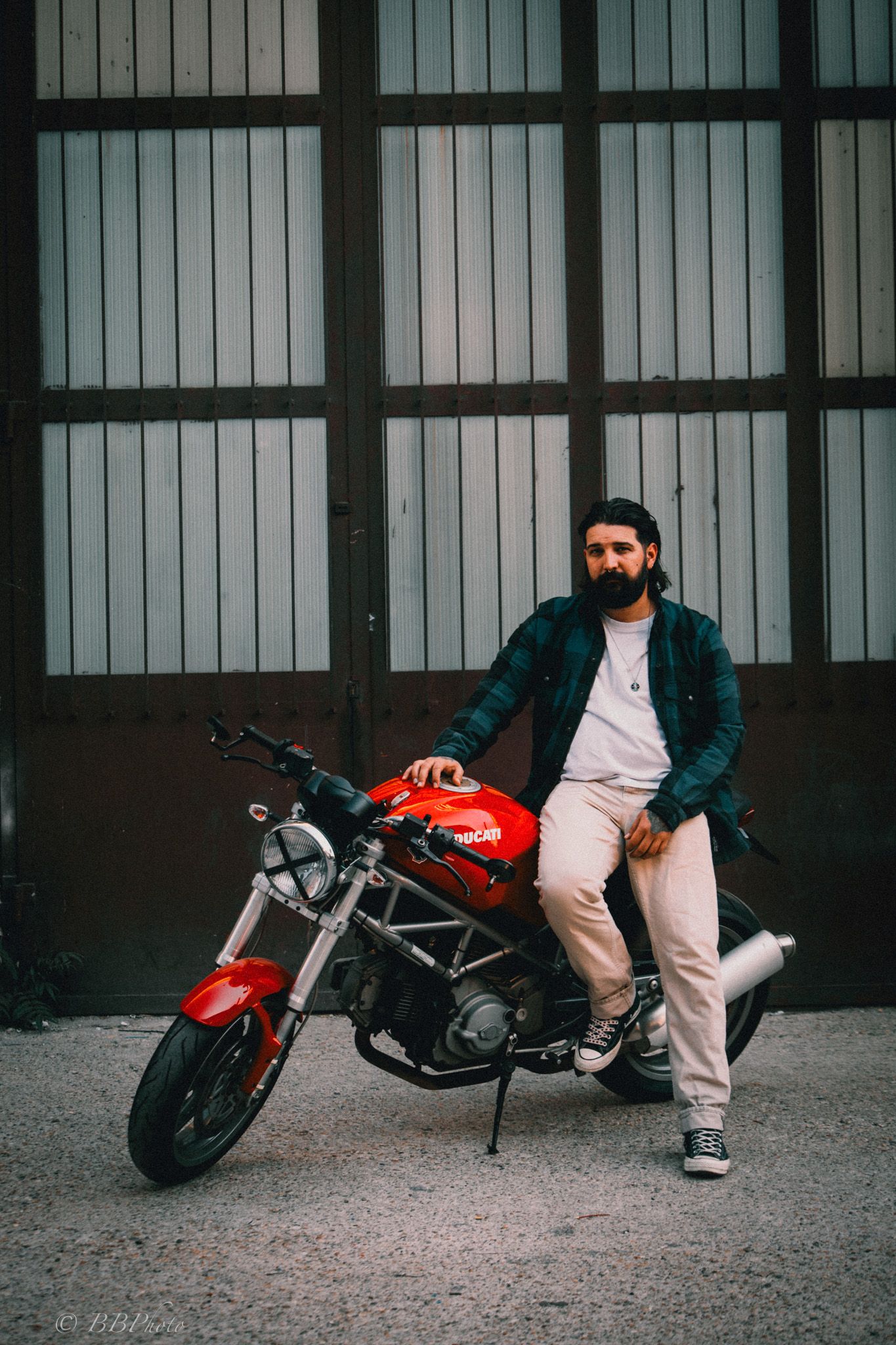 Bearded man in a plaid shirt and beige pants sits on a red naked motorcycle in front of a large industrial metal garage door.