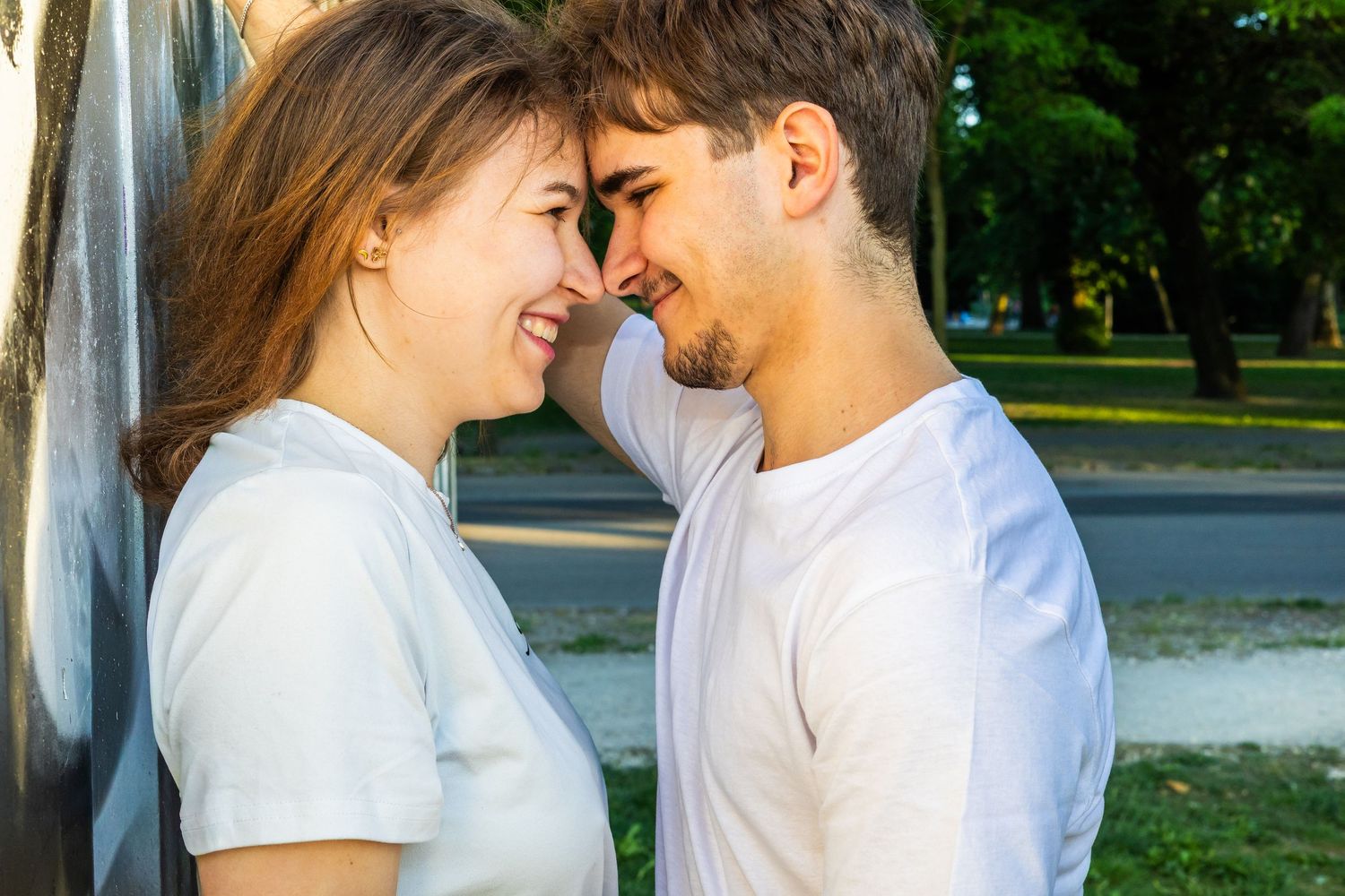 Young couple in white t-shirts sharing a romantic moment outdoors, touching foreheads and smiling in a park.