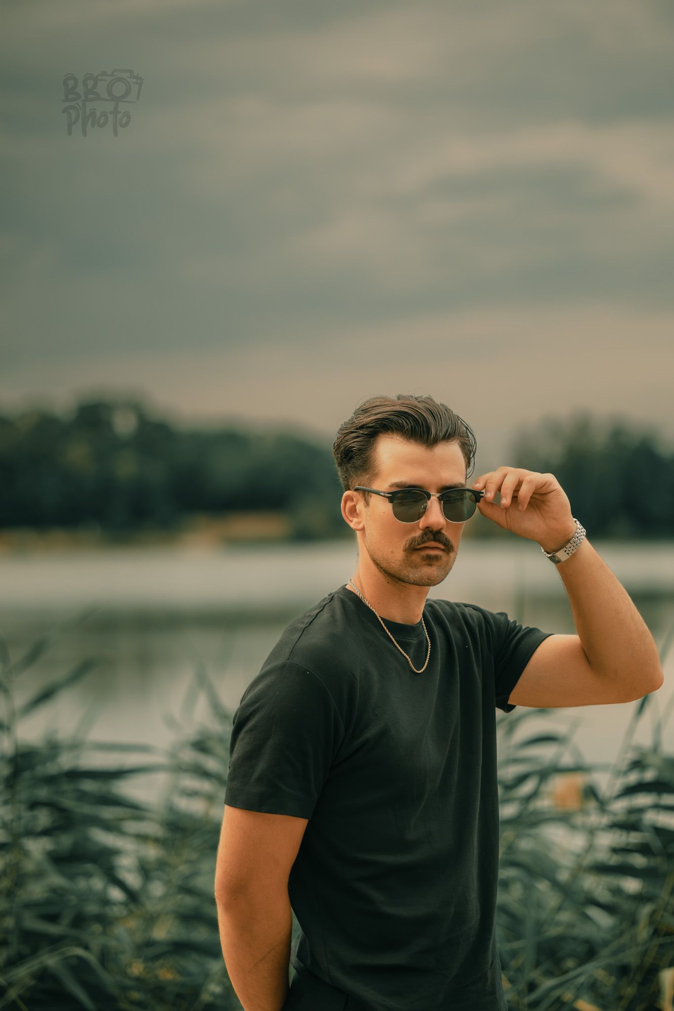 Stylish man with mustache adjusting sunglasses wearing a black t-shirt near a lake.