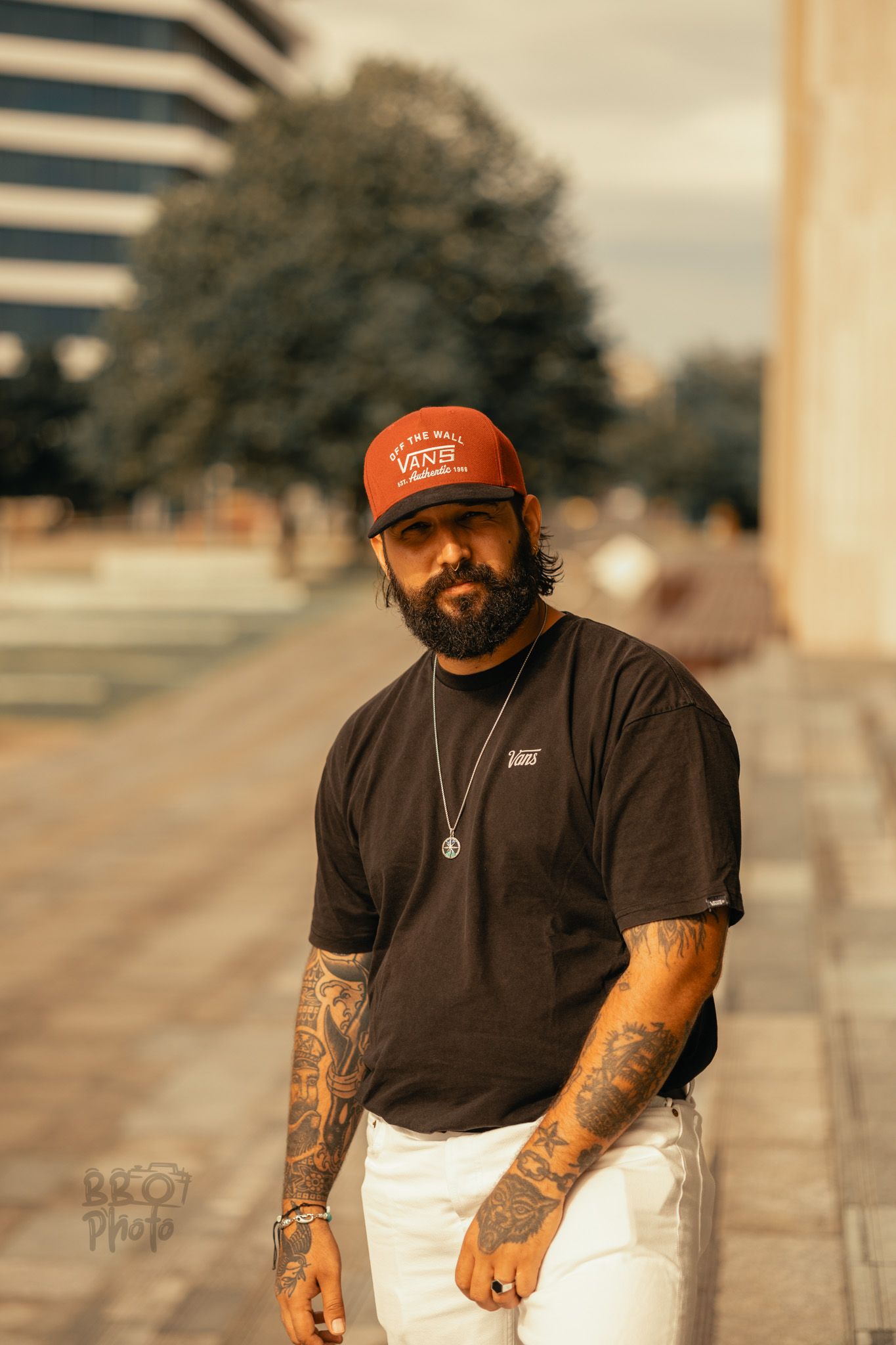 Tattooed man wearing a red Vans cap and black t-shirt poses outdoors on a sunny day.
