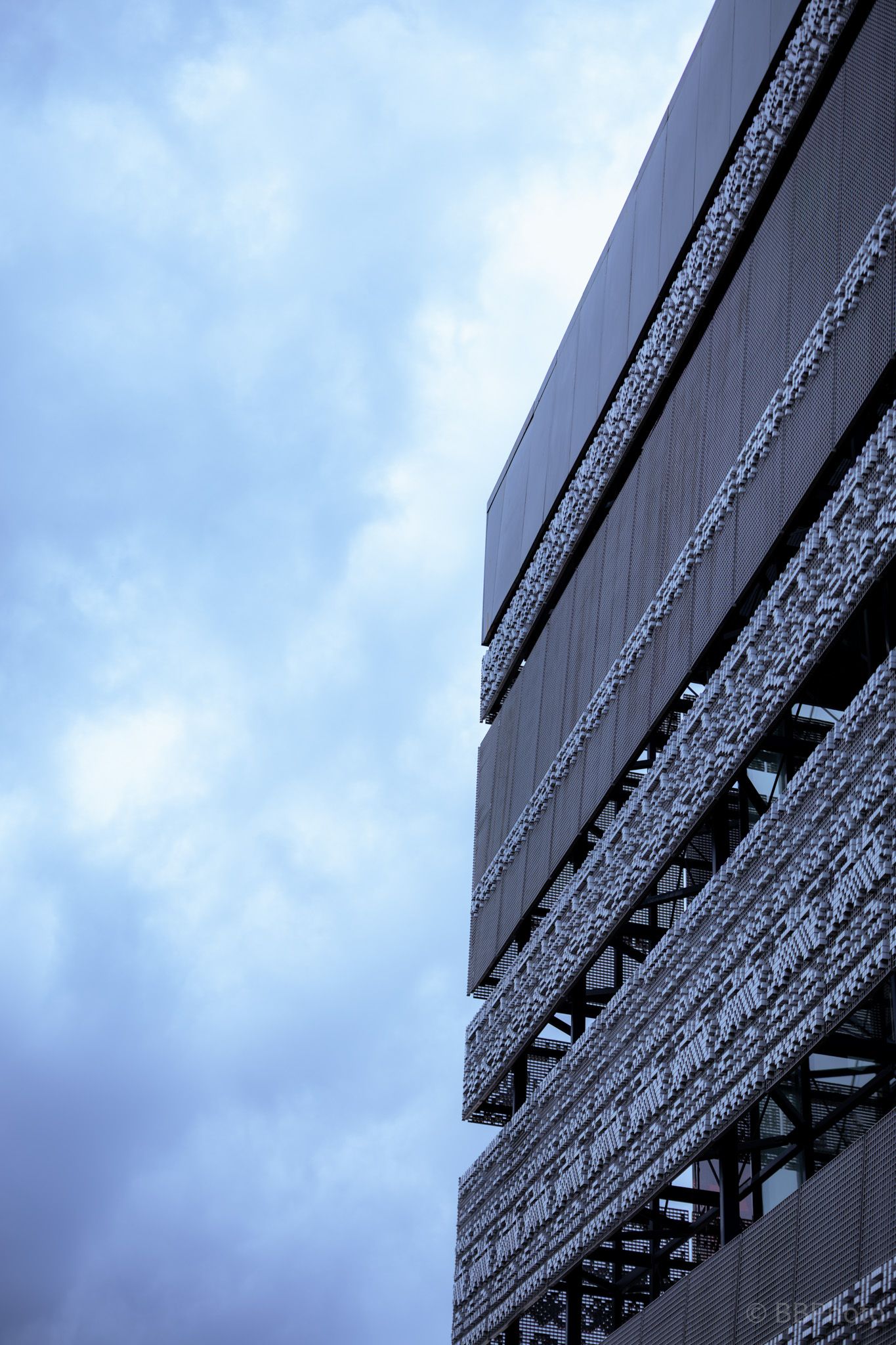 Low-angle view of a modern building facade with geometric metal panels against a dramatic cloudy blue sky.