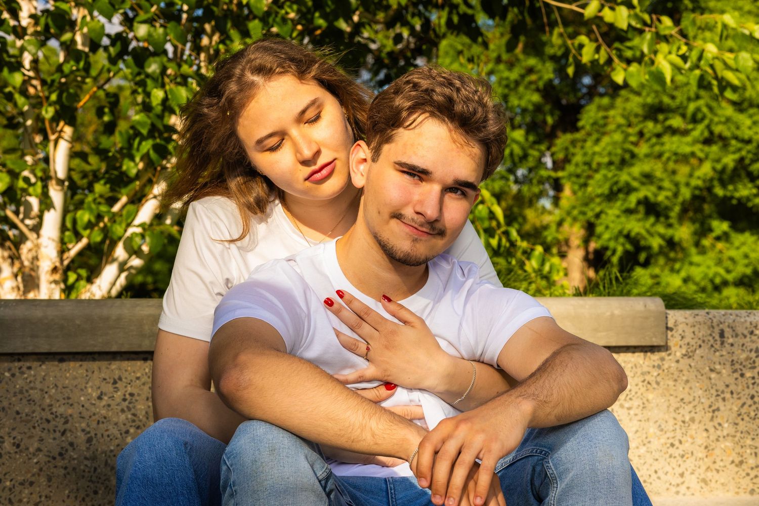 Young couple in white t-shirts sitting outdoors, woman resting head on man's shoulder showing engagement ring.