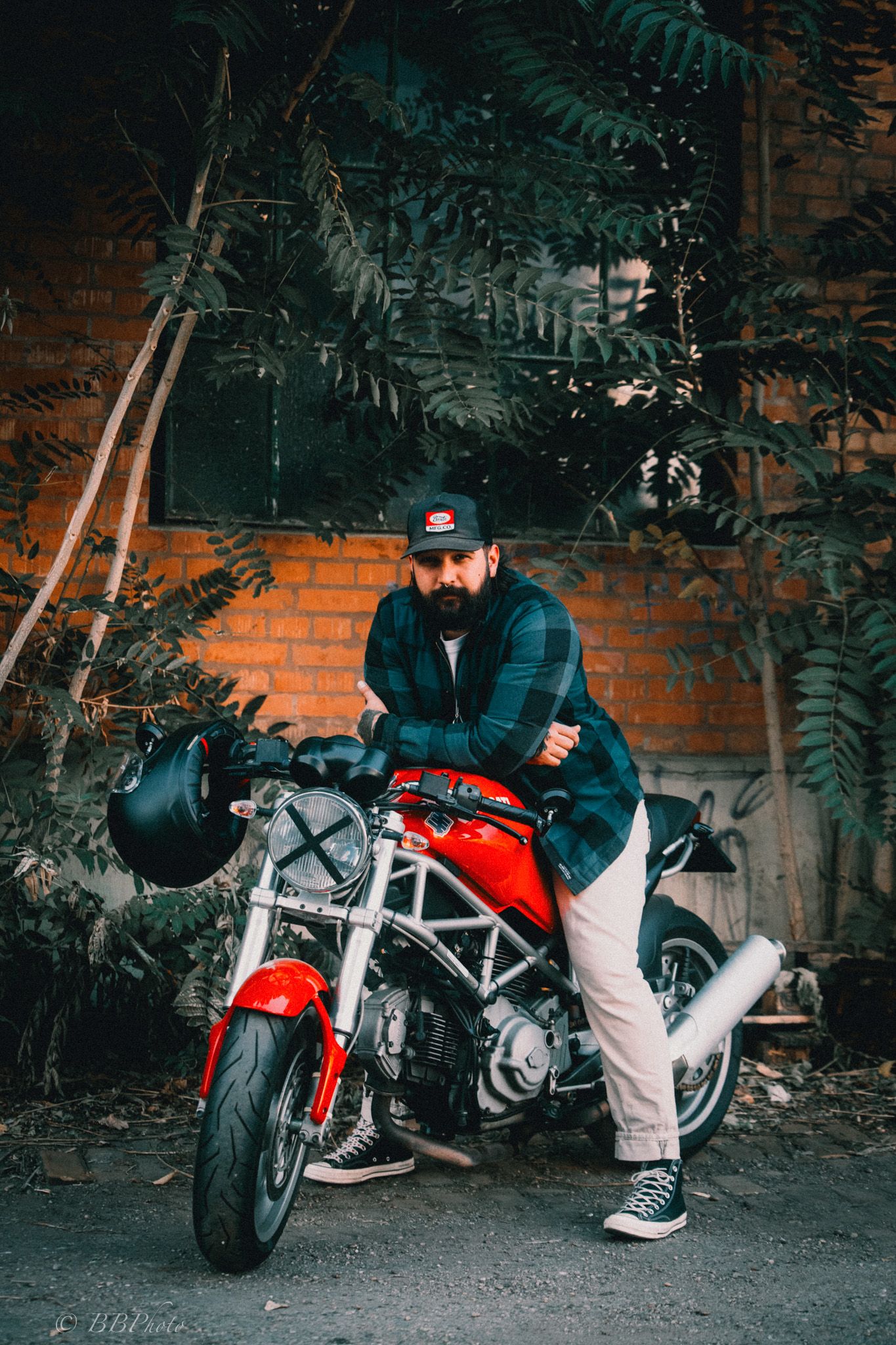 Bearded man in a plaid shirt and cap leans on a red and silver Ducati Monster motorcycle in front of a brick wall with green foliage.