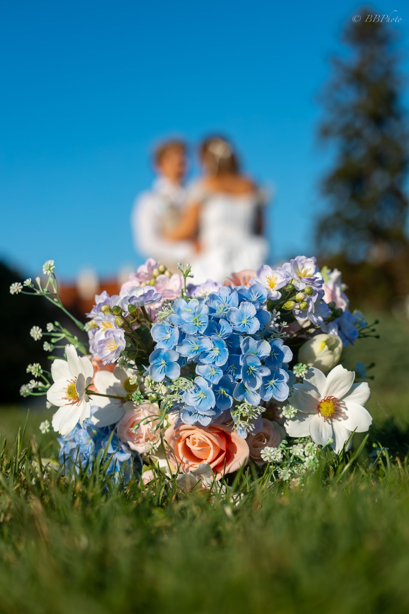 Wedding bouquet with blue, pink, and white flowers on grass, with blurred bride and groom embracing in background.