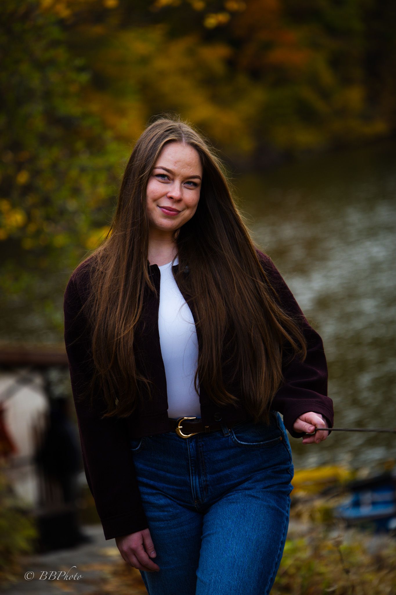 Smiling young woman with long brown hair in a dark jacket, white top and jeans standing outdoors by a lake with autumn foliage.