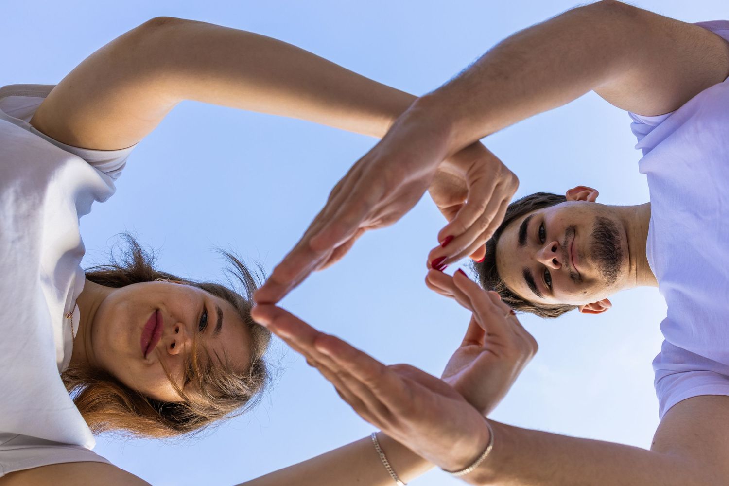 Two people in white shirts form a heart shape with their fingers against a blue sky, viewed from below.