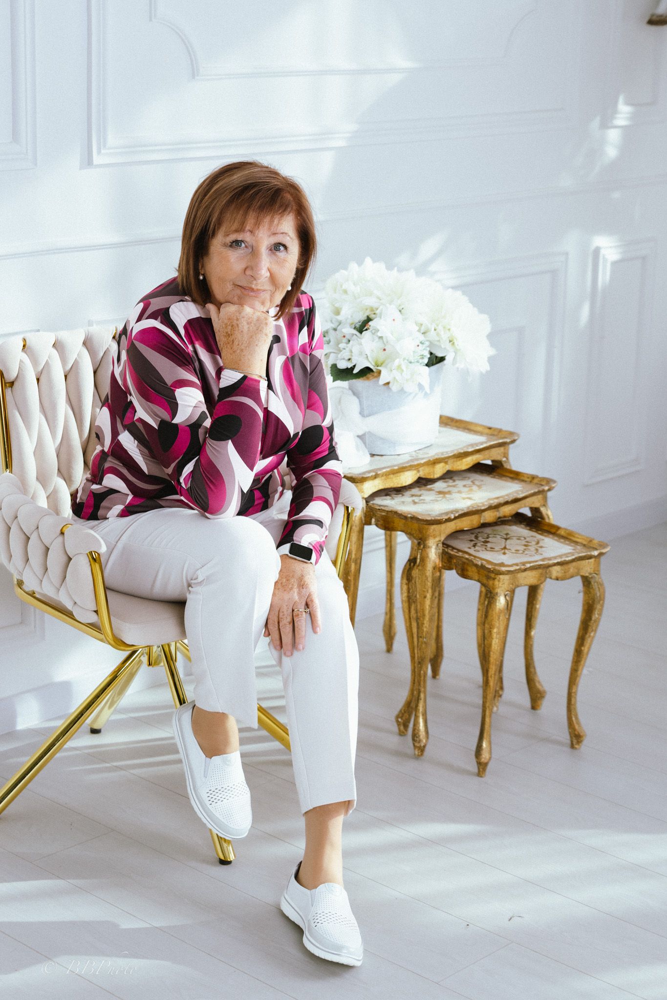 Smiling woman in a pink and burgundy patterned blouse and white pants sits on a gold chair beside gilded nesting tables and white flowers.