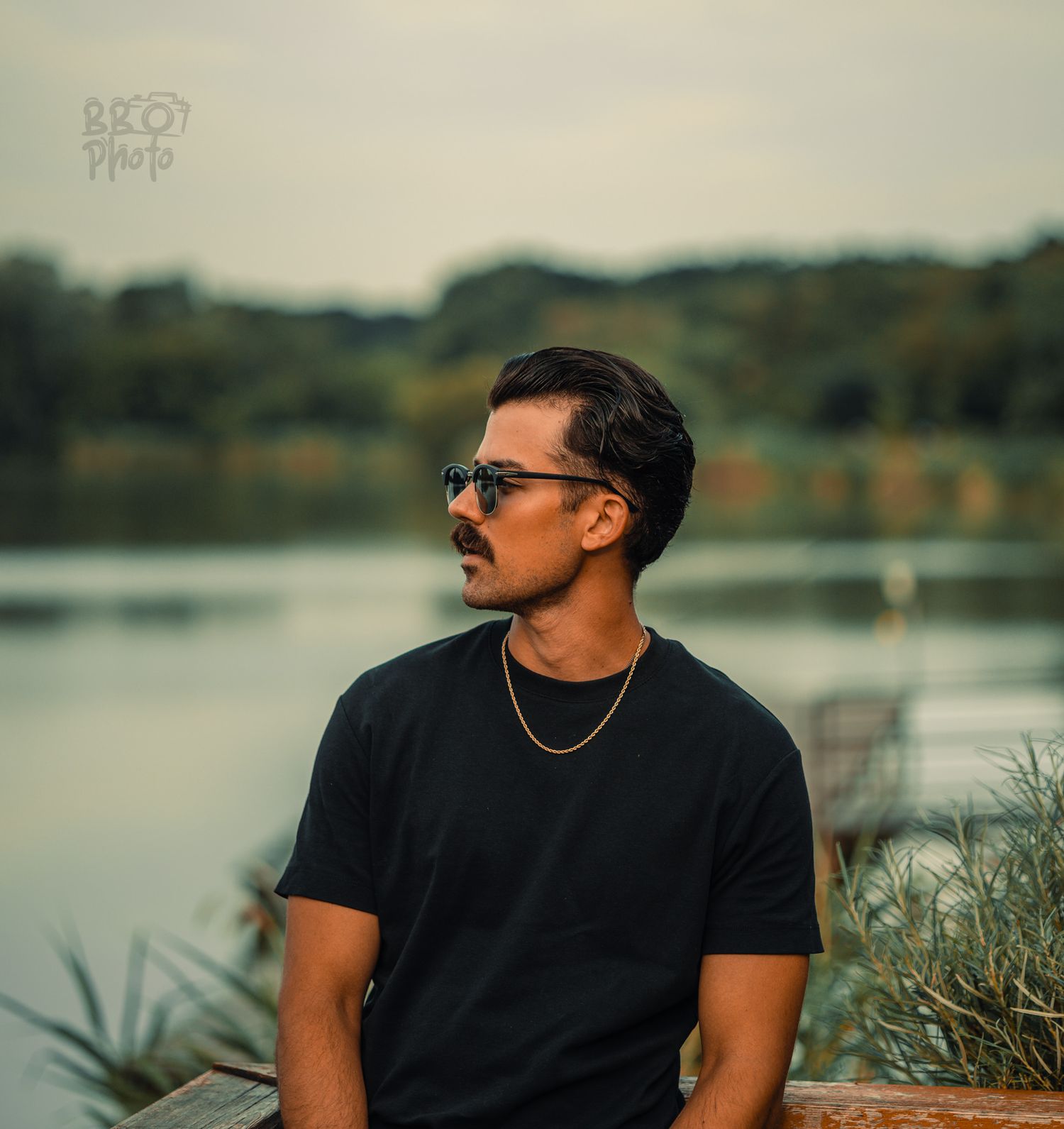 Man in black t-shirt and sunglasses sitting by a lake with lush green trees in the background.