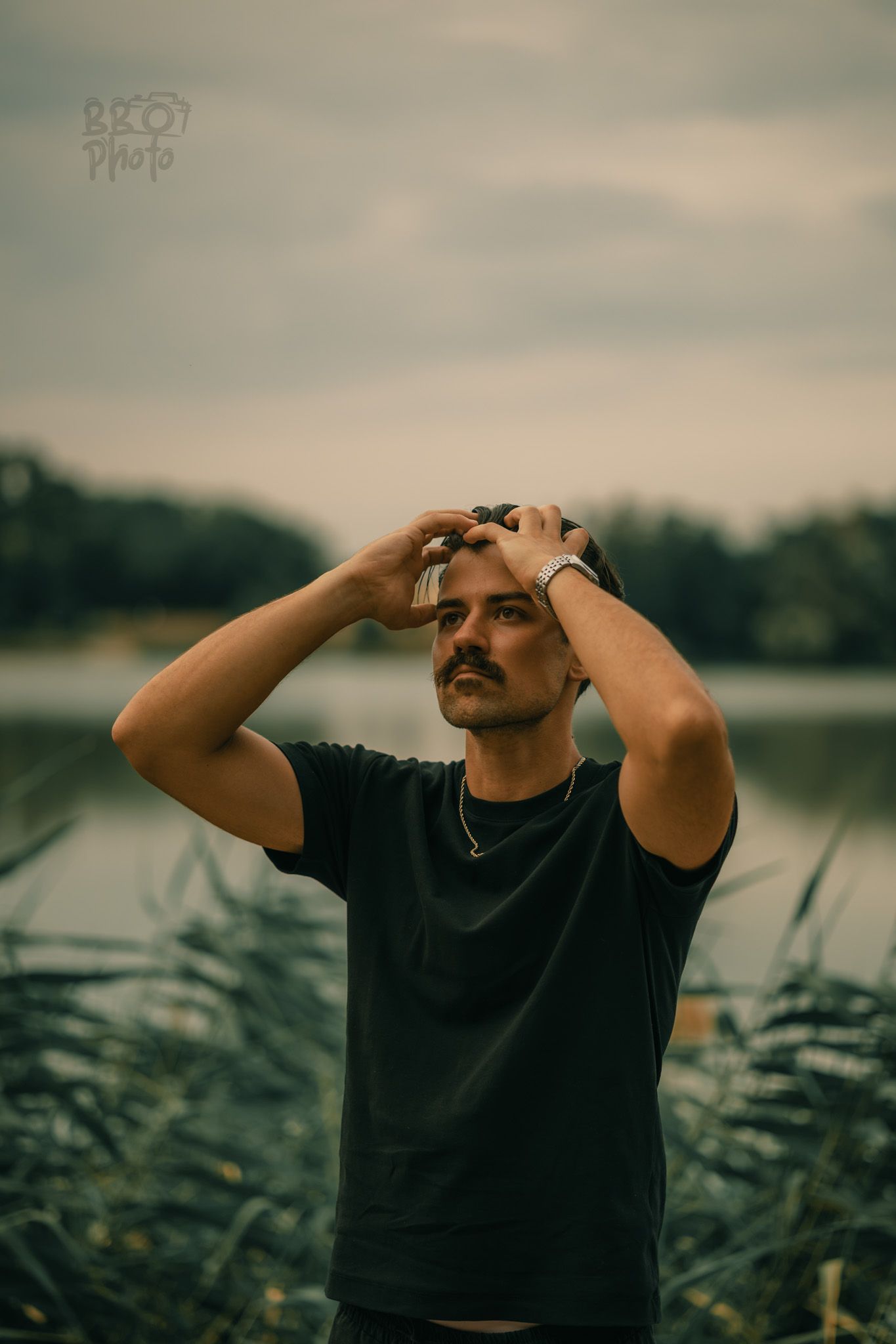 Man with mustache in black t-shirt holding his head with both hands standing near a lake.