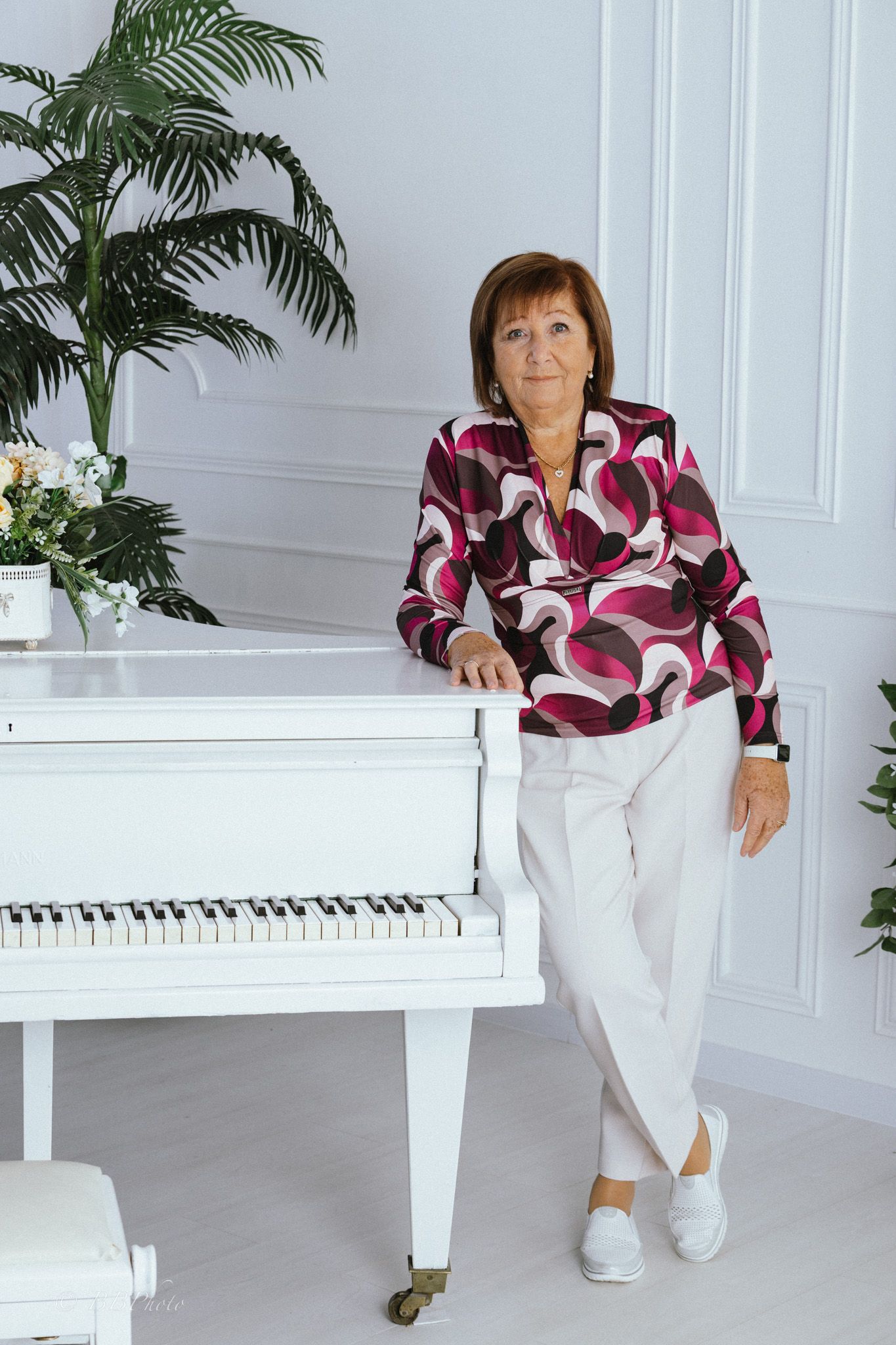 Smiling woman in a pink and burgundy patterned blouse and white pants leans on a white grand piano beside a potted palm in a bright room.